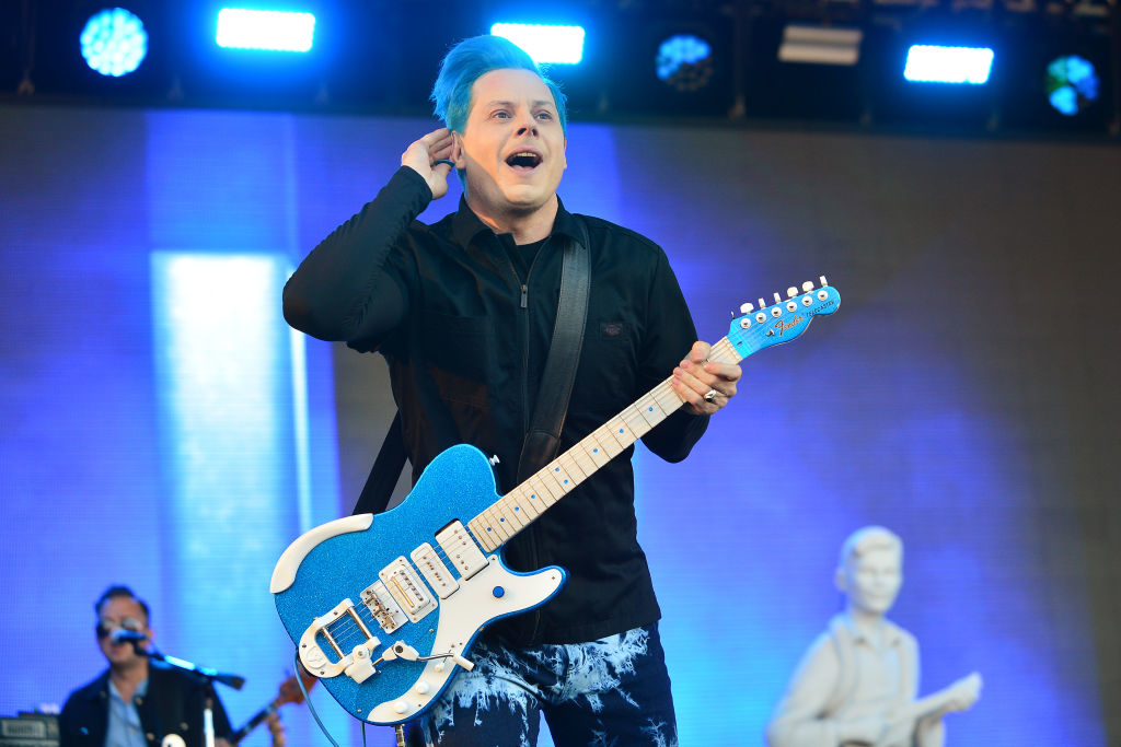 Jack White plays an electric guitar on stage, wearing a dark shirt and pants with a blue pattern, with blue hair and holding his ear