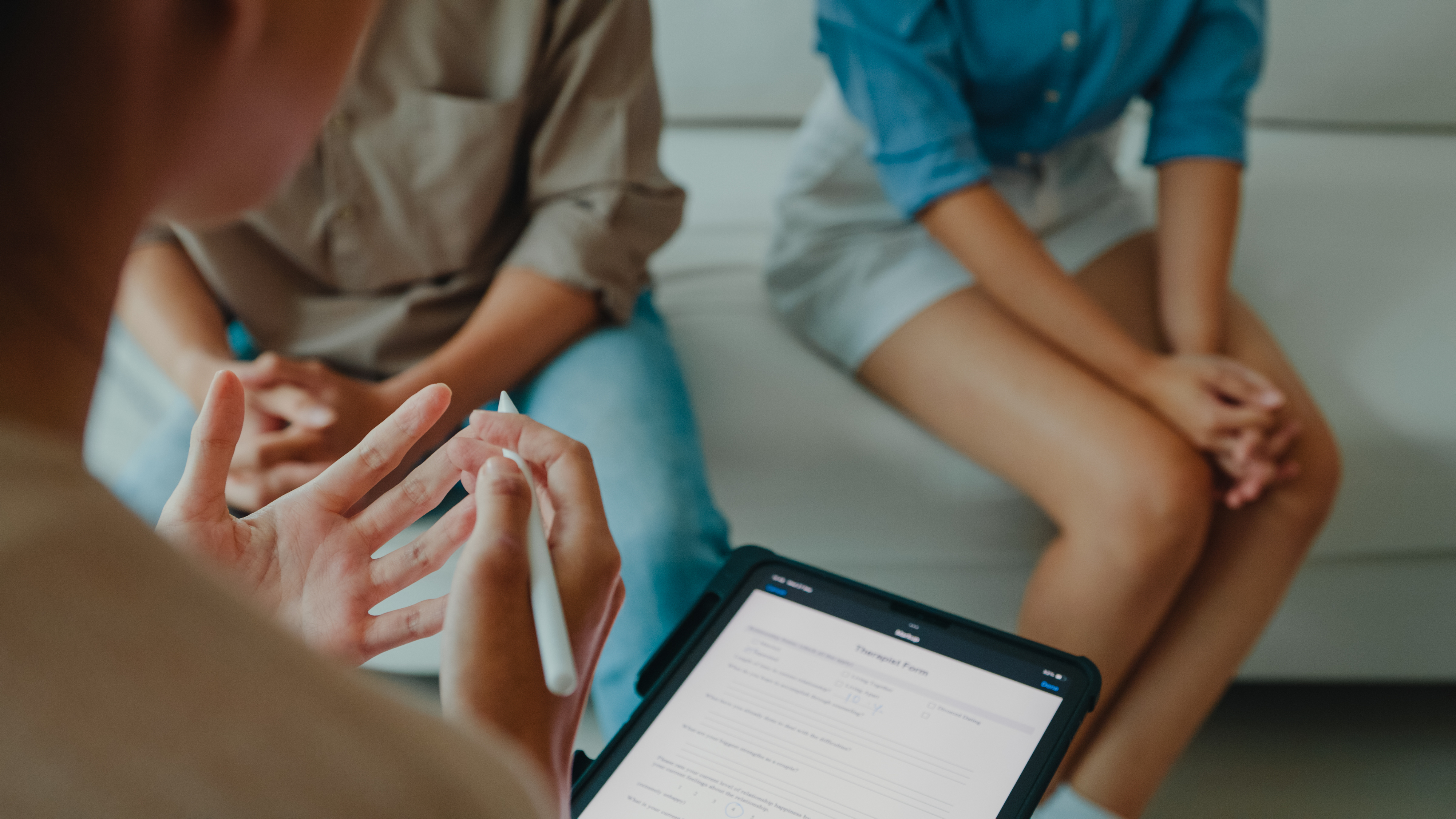 A couple sits on a couch during a counseling session, with a therapist holding a clipboard and taking notes. Names are not known