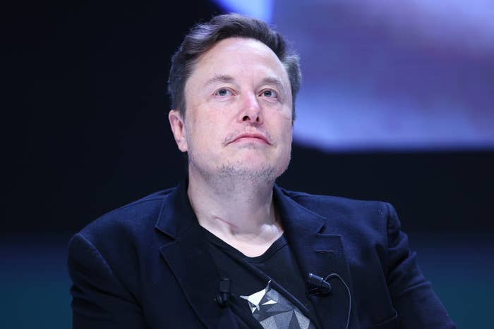 Elon Musk is seen slightly looking up, wearing a dark blazer over a graphic T-shirt, with a pensive expression in front of a blurred background
