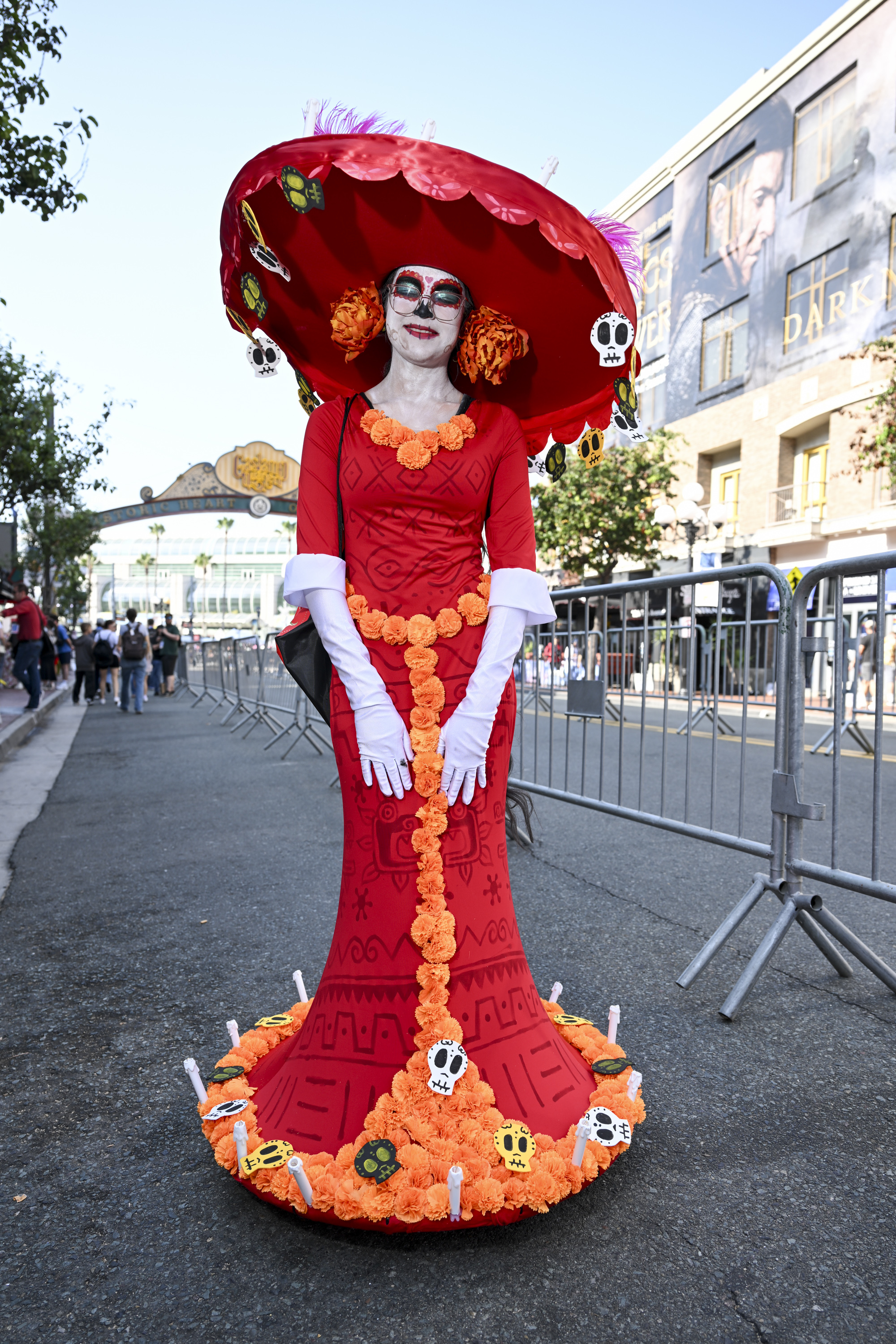 Person in an elaborate Day of the Dead costume with a large, wide-brimmed hat adorned with flowers and skulls, matching dress with candles, posing outdoors