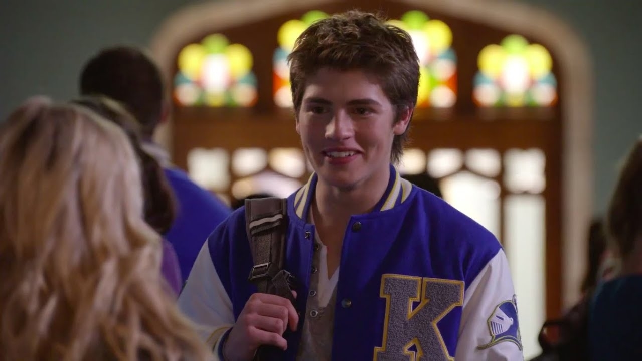 Gregg Sulkin wears a letterman jacket while talking to another person inside a building with stained glass windows in the background