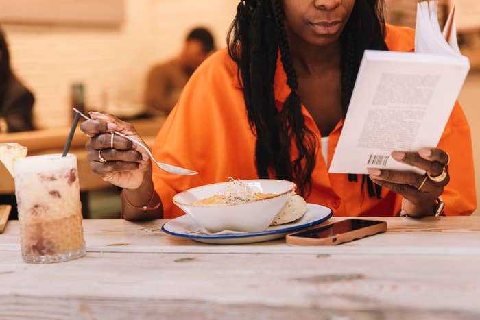 A person in an orange shirt reads a book while eating a meal at a wooden table. A smartphone and a drink with ice are beside the meal