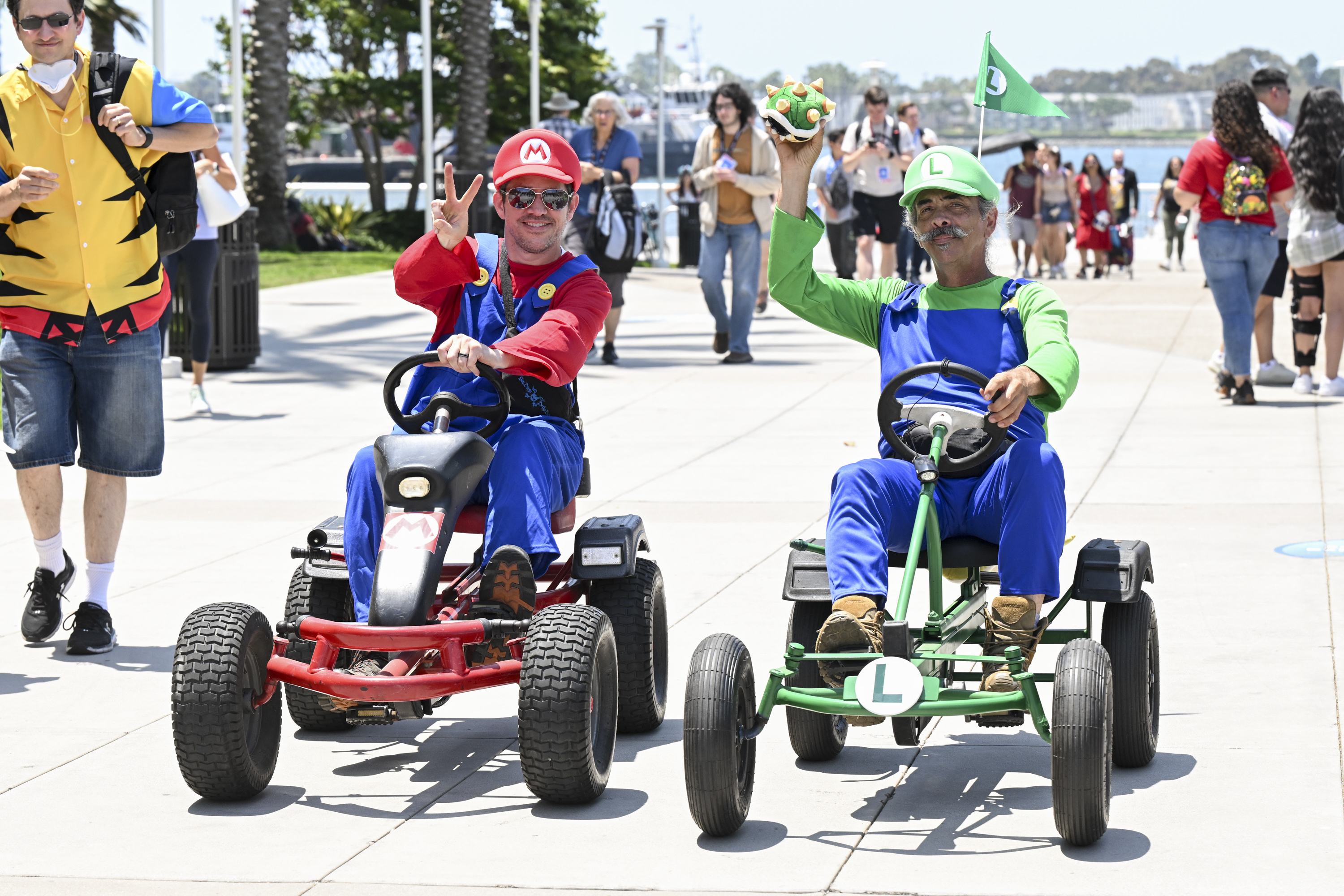 People dressed as Mario and Luigi from the Super Mario series ride go-karts outdoors, making victory signs and smiling. Passersby and trees in the background
