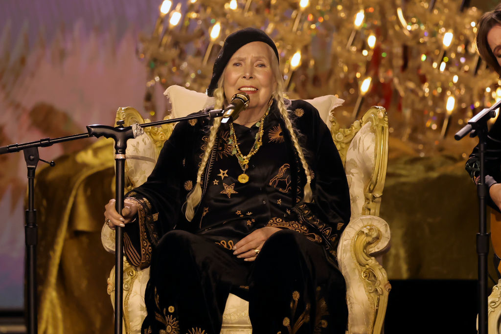 Joni Mitchell performs while seated on stage, wearing a black and gold outfit with star patterns, against a backdrop of a golden decorative display