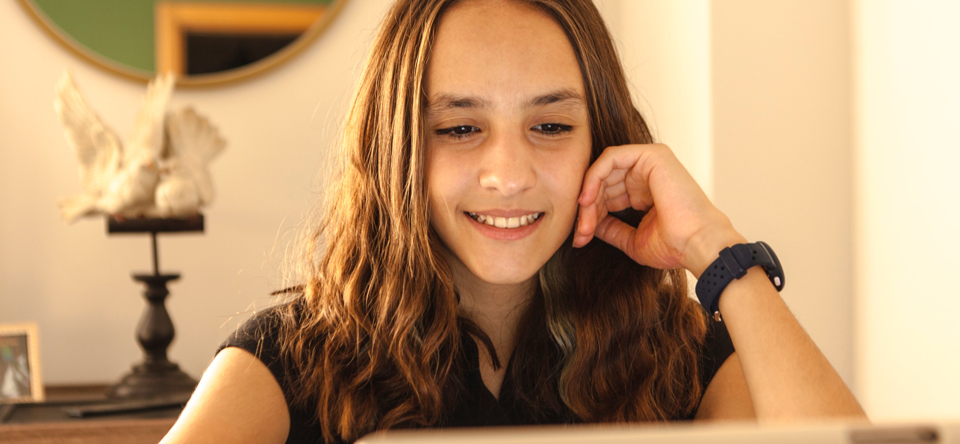 A smiling girl with long hair uses a tablet while sitting at a desk with family photos and a circular mirror in the background