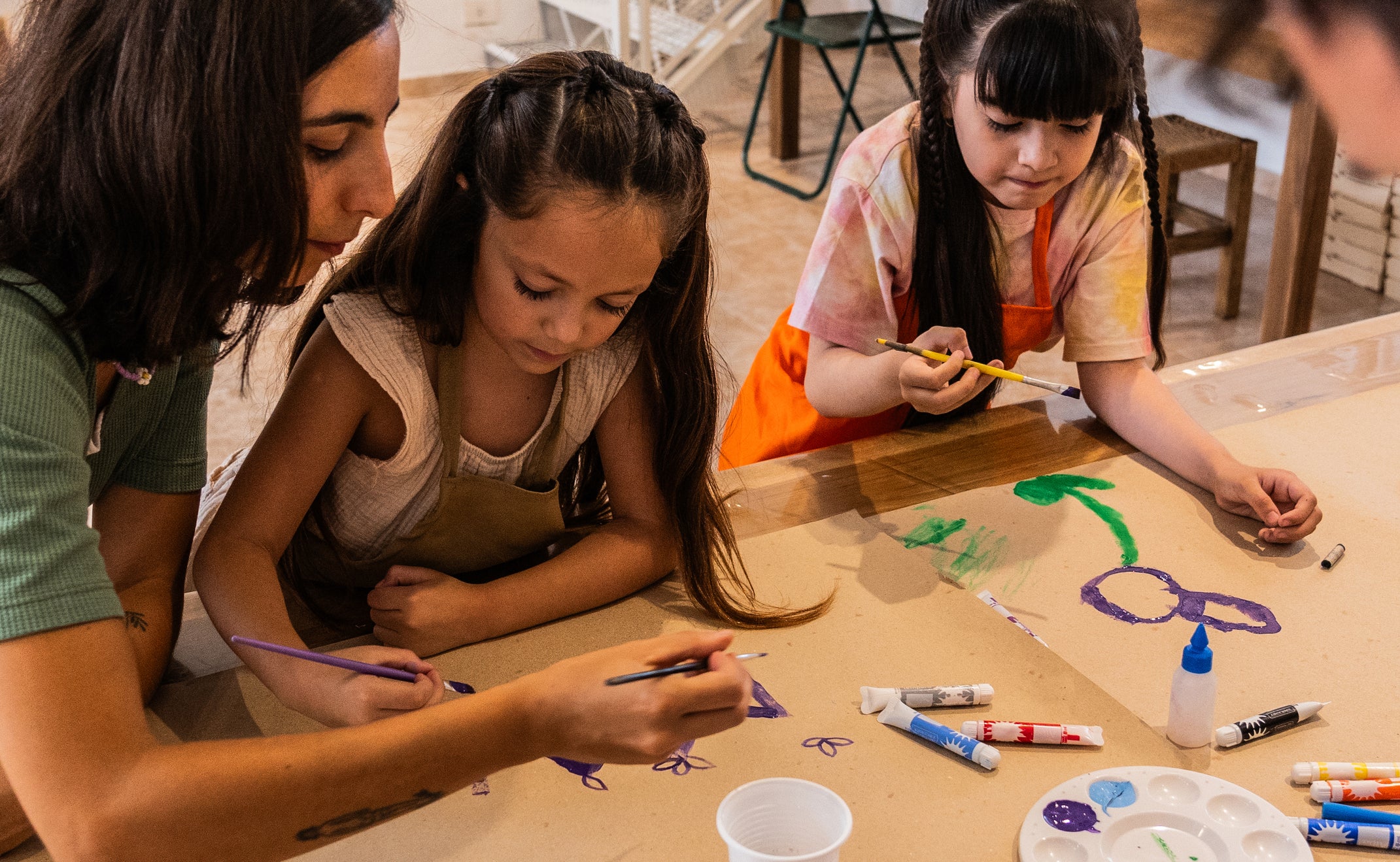 A woman and three children, including two girls, are engaged in painting and drawing activities at a table covered with art supplies