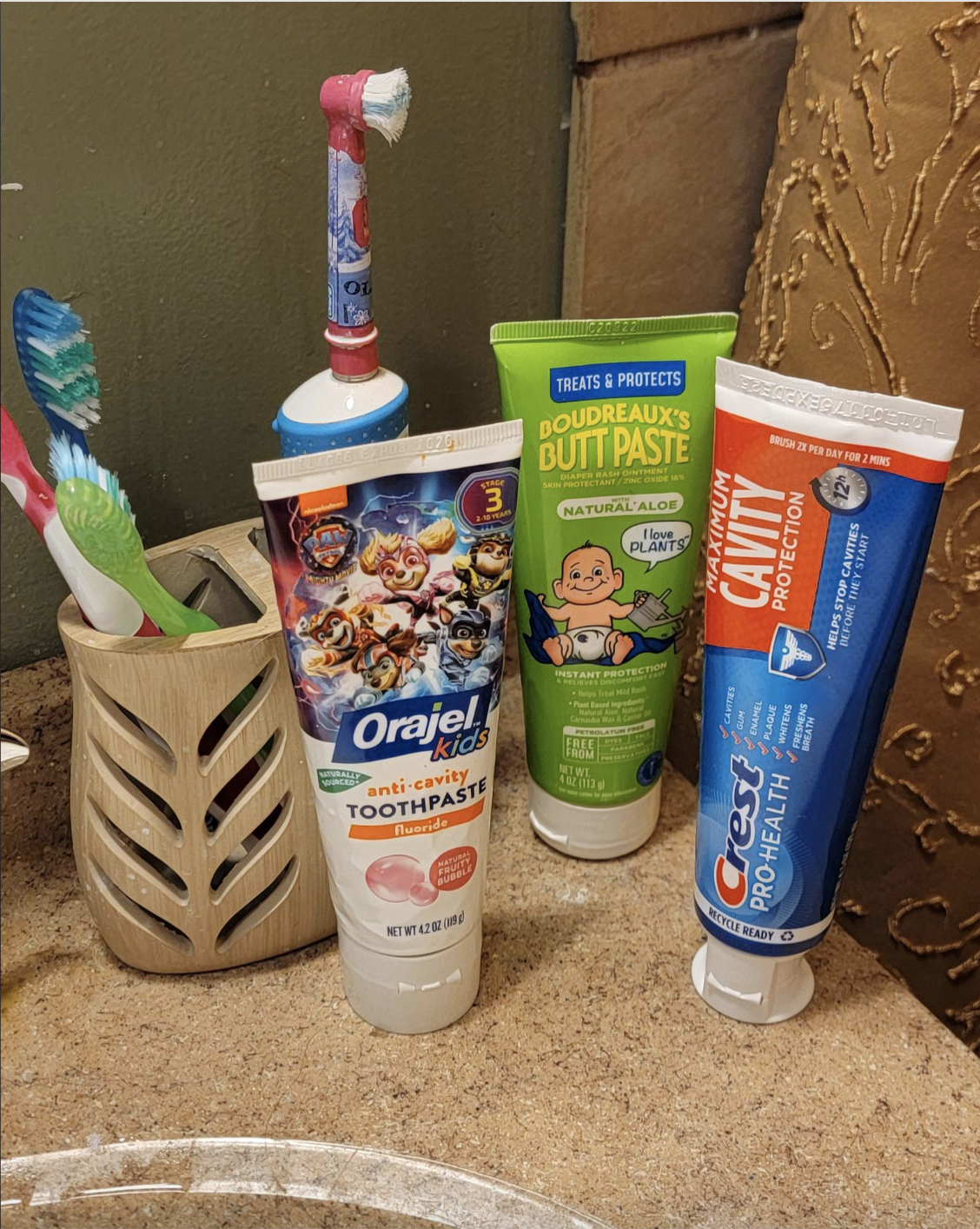 Toothbrushes and three toothpaste tubes, including Orajel Kids Toothpaste, Boudreaux's Butt Paste, and Crest Cavity Protection, on a bathroom sink