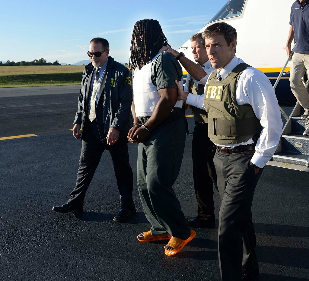 FBI agents escort a handcuffed individual with dreadlocks, wearing prison attire and orange sandals, off an airplane on a tarmac