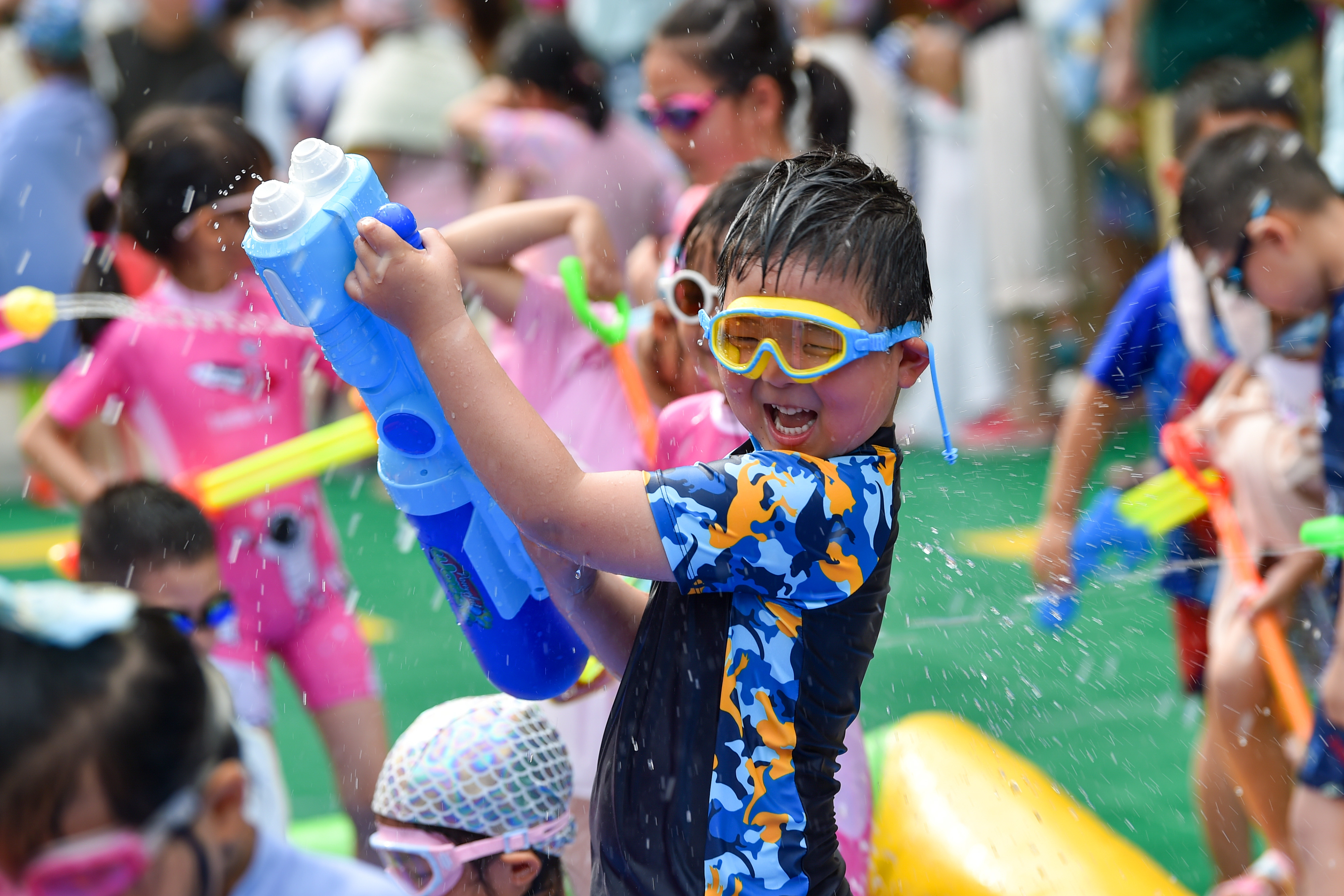 A child in a swim shirt and goggles joyfully sprays water from a large toy gun, surrounded by other kids playing with water toys at a crowded event