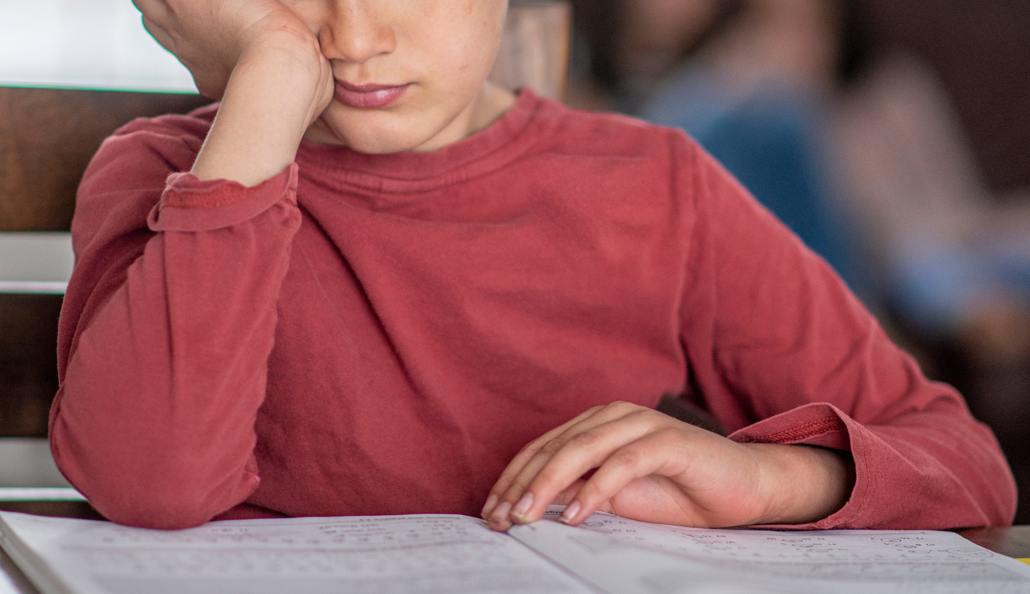 A young boy, wearing a long-sleeve shirt, looks thoughtfully at a book he is reading. Two adults are sitting on a couch in the background