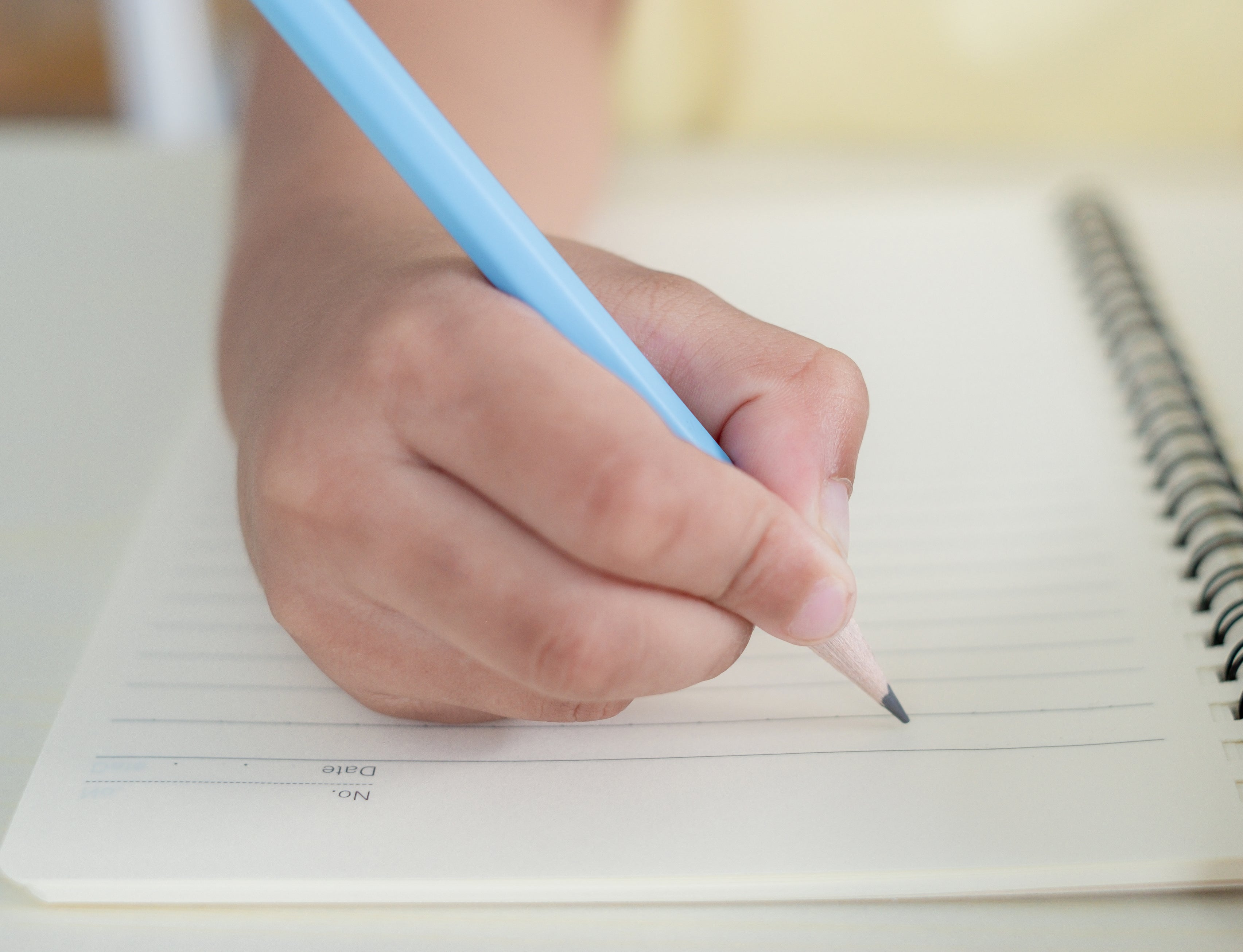 A hand holding a blue pencil writes in a spiral-bound notebook with lined pages