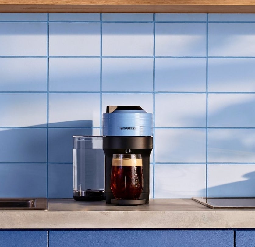 A kitchen counter with a Nespresso machine brewing coffee. The background includes tiled walls, cookbooks, and glass cups on a shelf