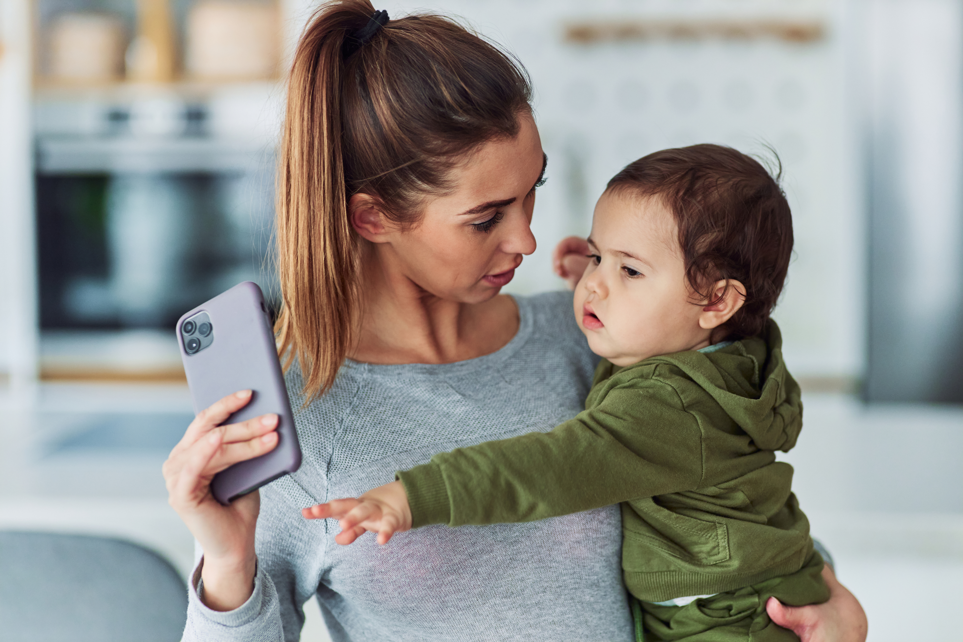 A woman in casual clothing holds a smartphone with one hand and carries a toddler in a green hoodie with the other