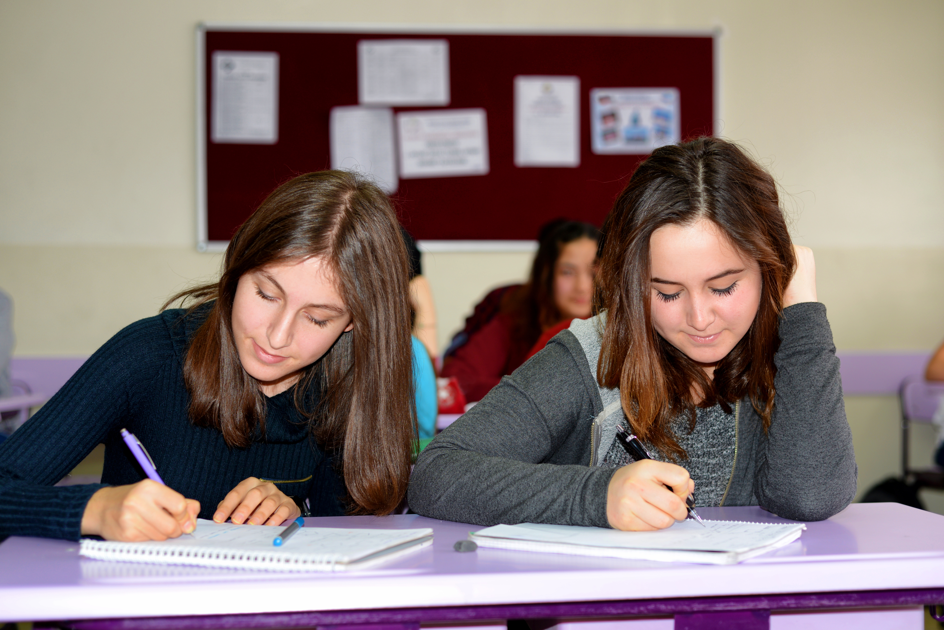 Two teenage girls are sitting at desks in a classroom, focused on writing in their notebooks. A bulletin board with papers is visible in the background