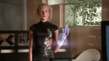 A woman stands indoors, wearing a black top with floral embroidery and holding a holographic interface