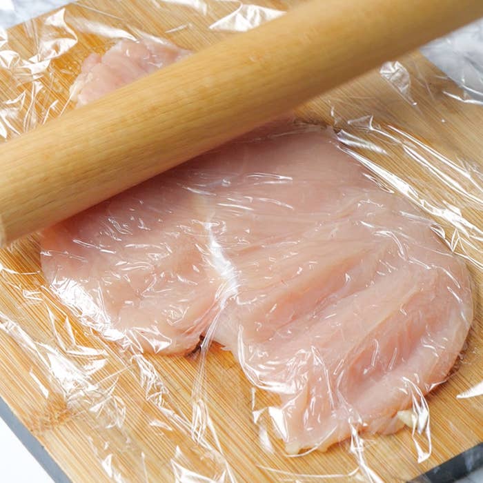 まな板の上に置かれたラップで覆われた鶏肉を、麺棒で平らに伸ばしている様子。