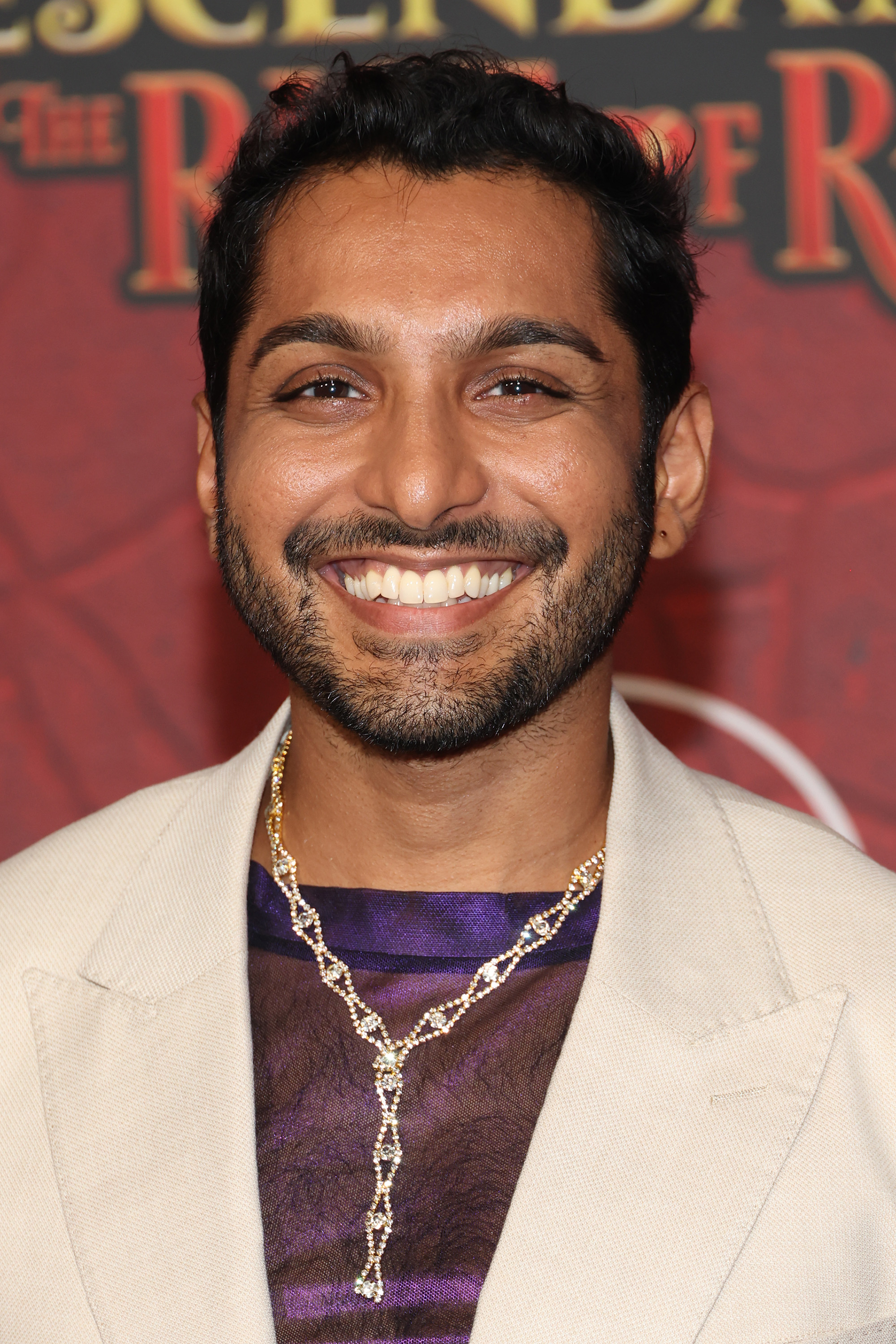 Levin Valayil on the red carpet for "Descendants: The Rise of Red" in a cream jacket, purple shirt, and ornate necklace