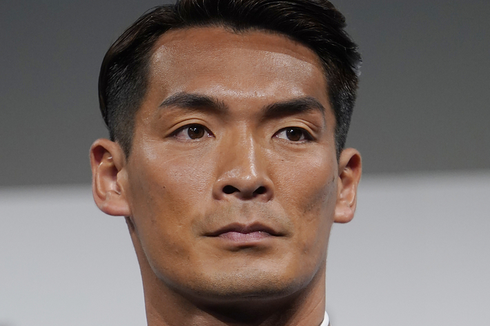 男性が茶色のスーツとストライプのシャツ、黒いネクタイを着ている。