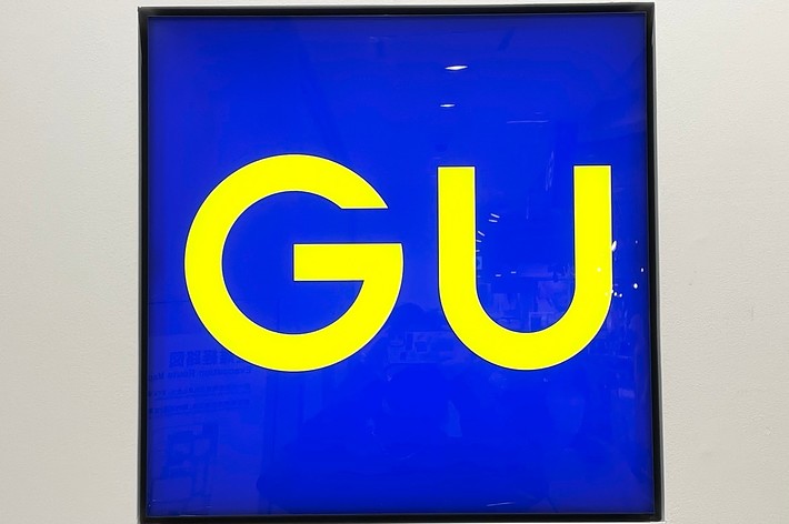 Square blue sign with bright yellow letters “GU” in a minimalist design