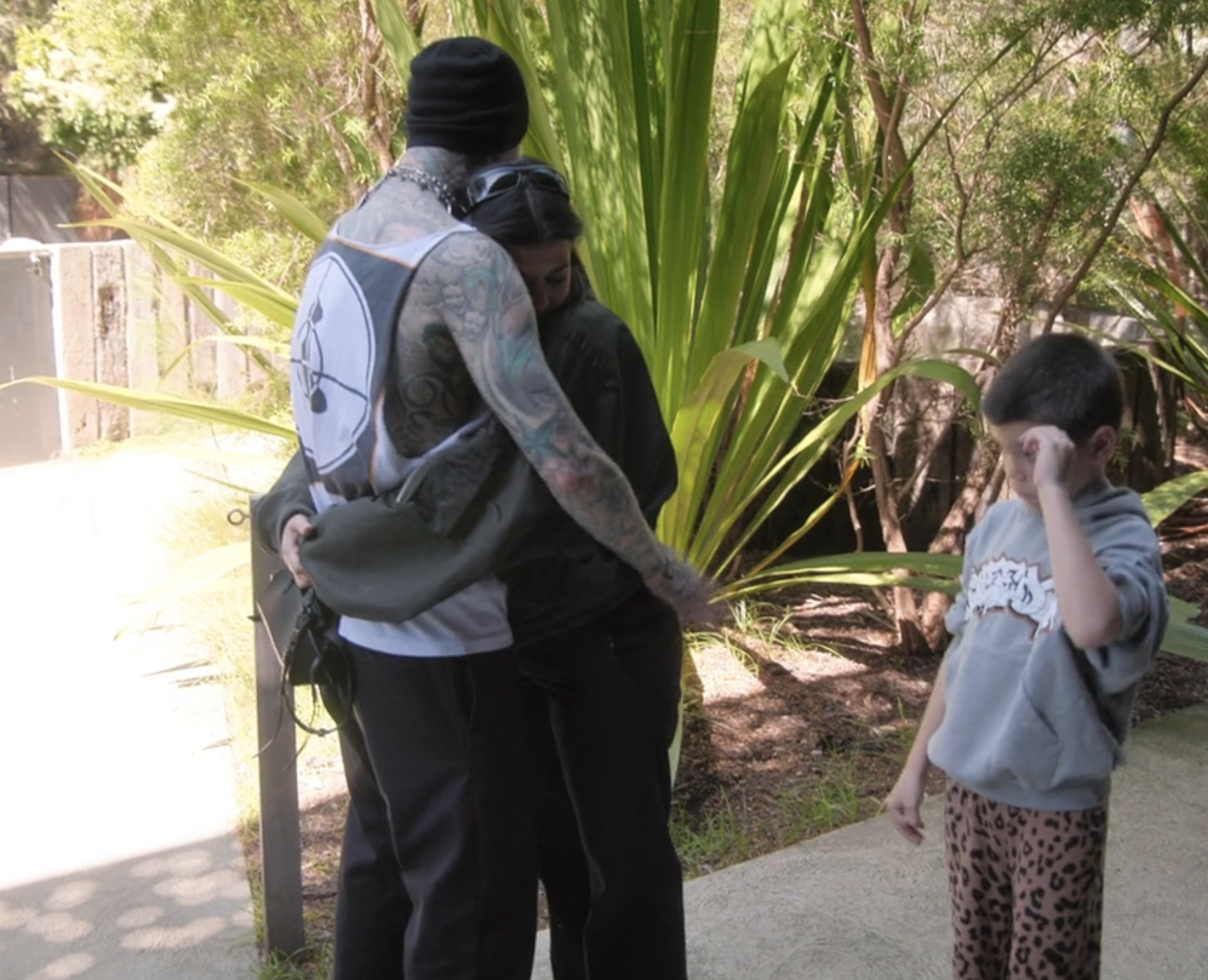 A man with tattoos and a beanie hugs a woman with dark hair while another woman, standing nearby, wipes her face