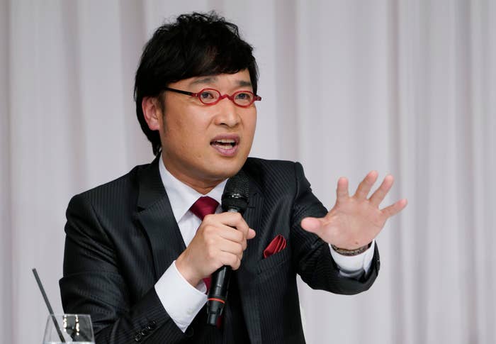 メガネをかけたスーツ姿の男性、手にマイクを持って話している。