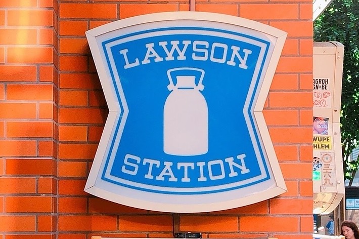 ローソンの24時間営業看板がレンガの壁に取り付けられている。背景に歩道と人々が見える。