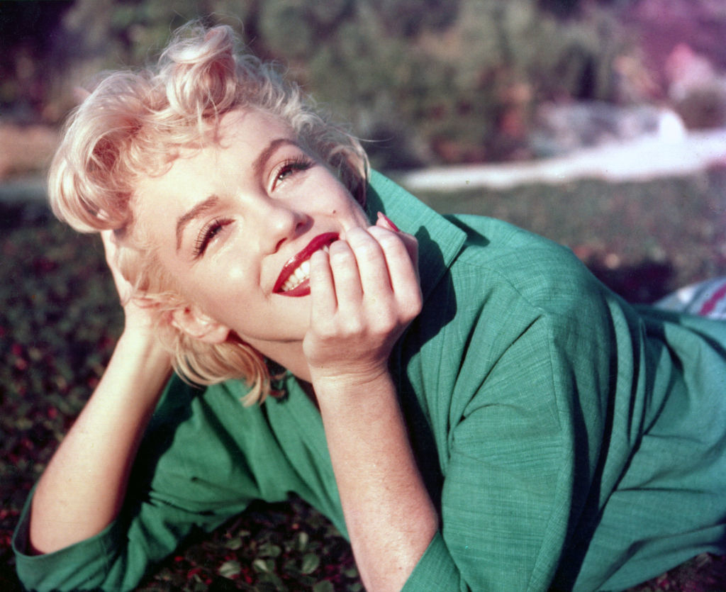 Marilyn Monroe smiles, lying on the grass with her head propped on one hand, wearing a green blouse