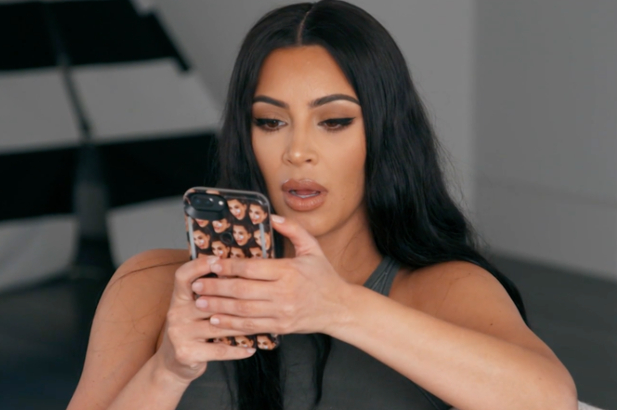 Kim Kardashian looking at her phone with a surprised expression. The phone case has multiple images of Kim Kardashian's face printed on it