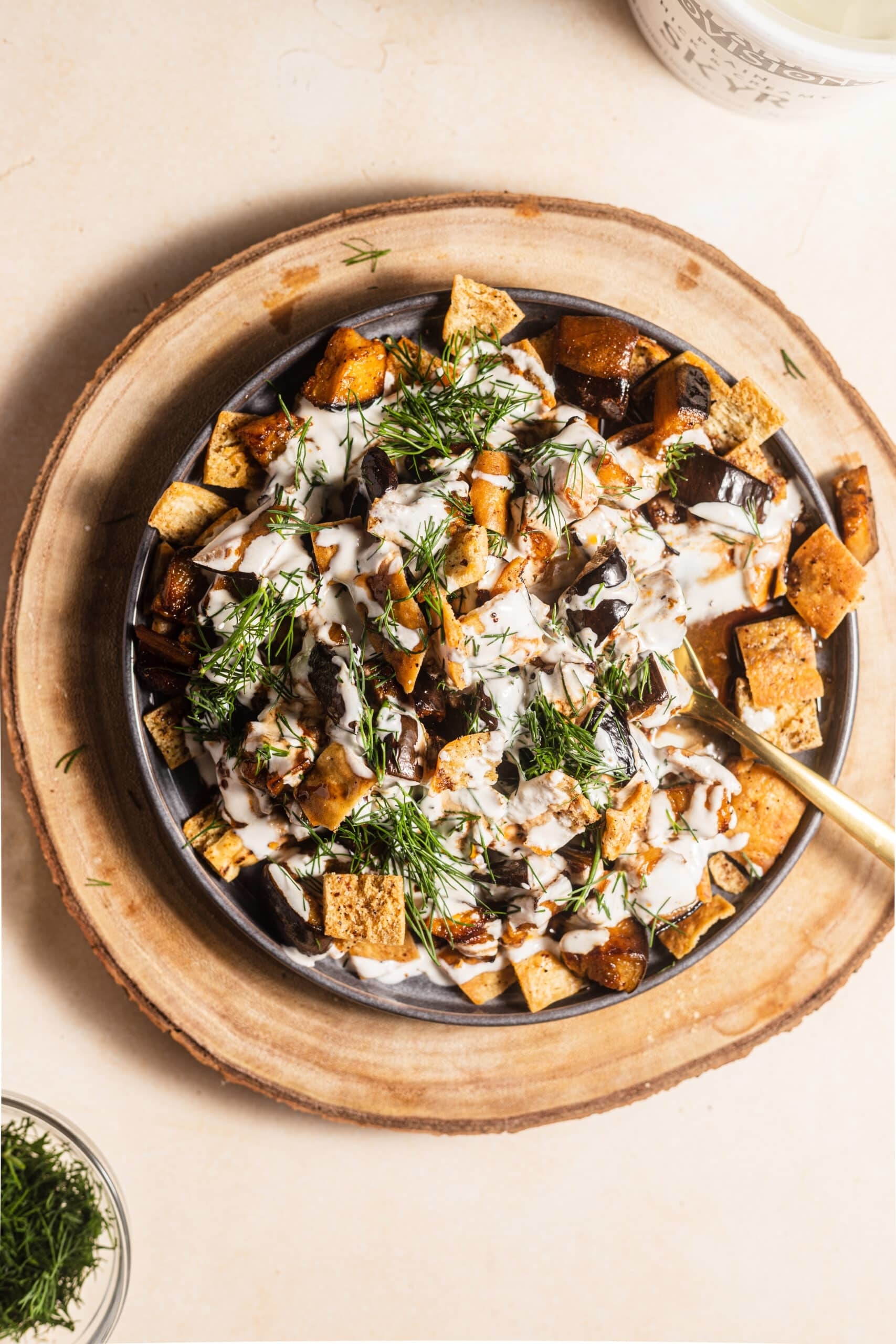 A plate of roasted vegetables topped with a creamy white sauce and fresh dill, with a golden fork on the side