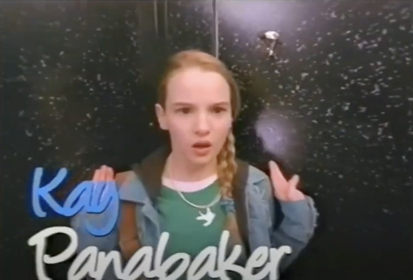 Kay Panabaker, wearing a casual outfit with a green shirt and denim jacket, stands against a speckled black background, appearing surprised or concerned