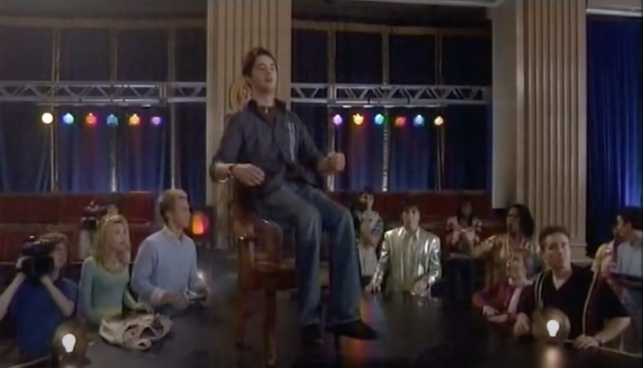 Group of people, including a person in a chair, film a scene in a large room with columns and colorful lights in the background