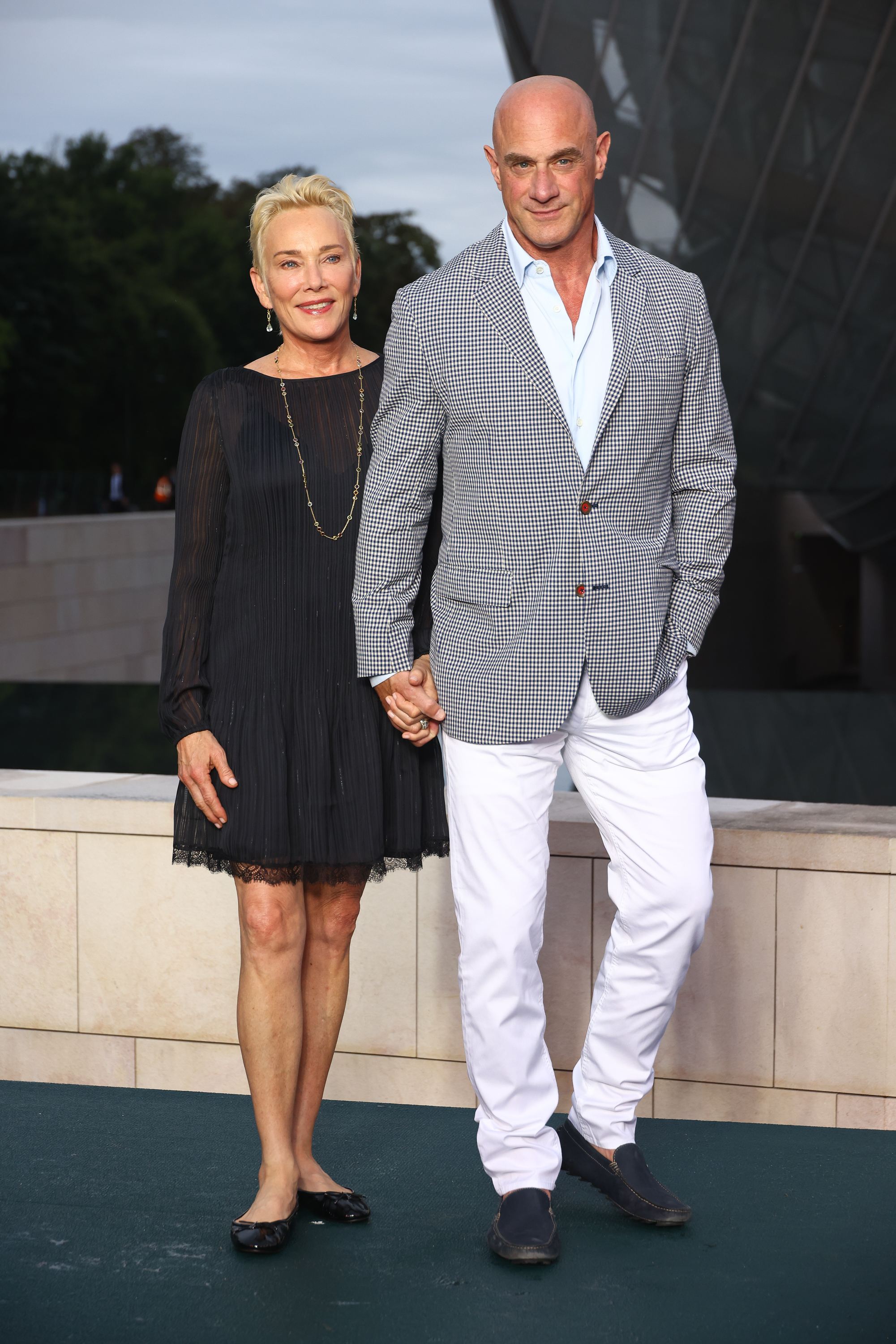 Sherman Williams and Christopher Meloni stand on a ledge holding hands. Sharon wears a chic black dress, and Christopher wears a checked blazer with white pants