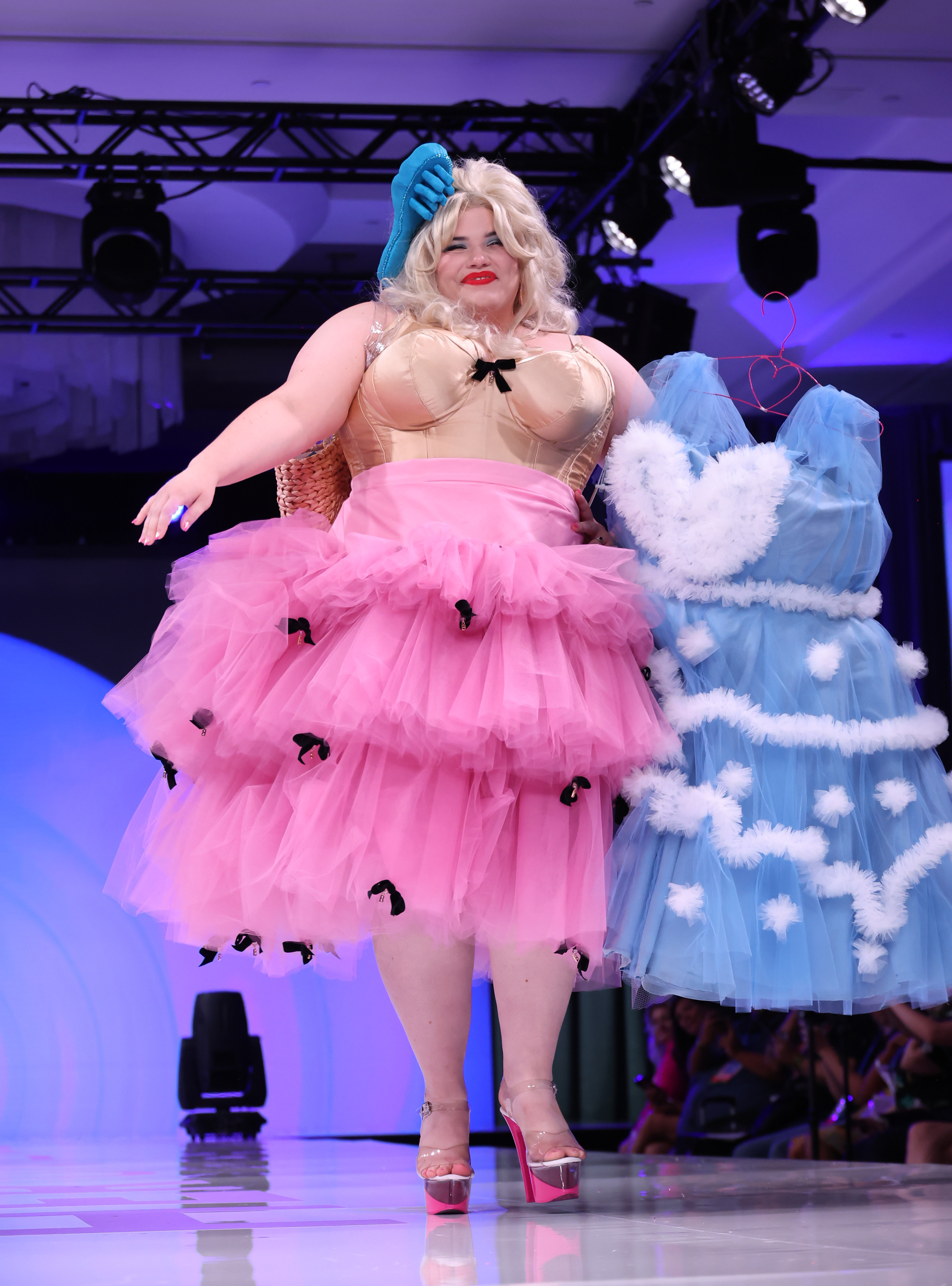 Woman in a layered pink tulle dress and blue headpiece holding a blue, fluffy, tulle dress on a runway