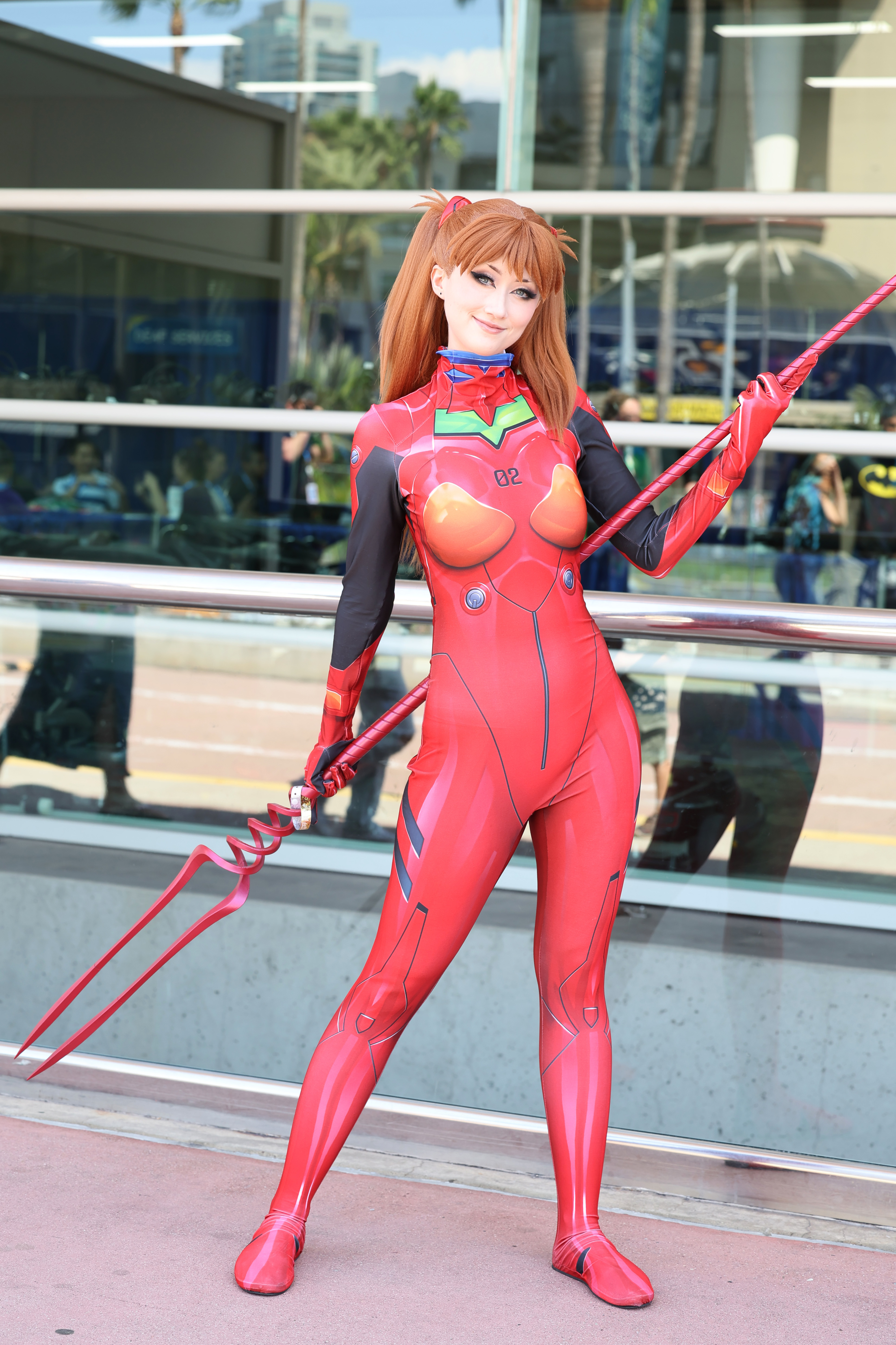 Cosplayer dressed as Asuka Langley Soryu from "Neon Genesis Evangelion" in a red unitard, holding a prop lance, poses outside a convention center