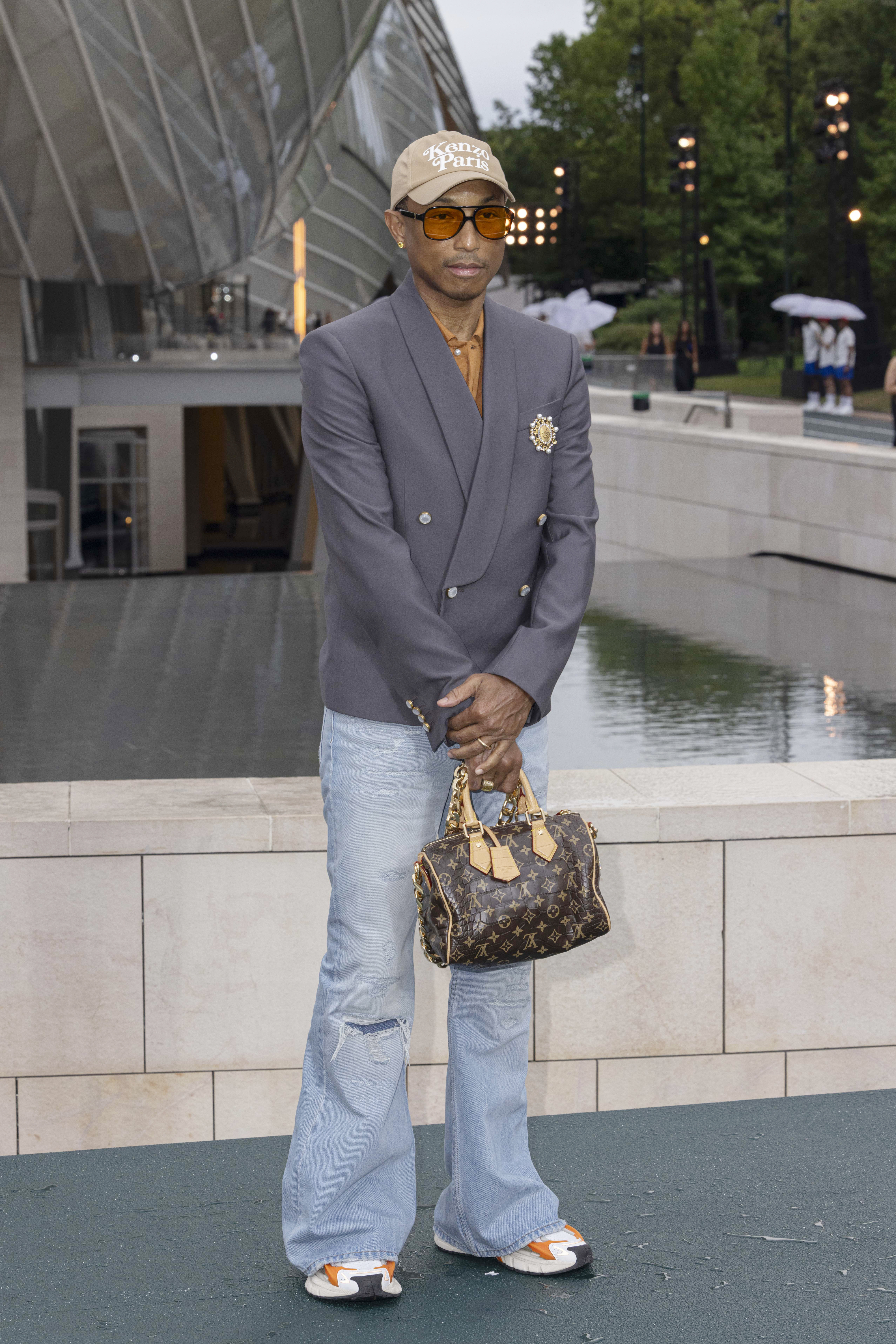 Pharrell Williams stands outdoors, wearing an oversized blazer, ripped jeans, a beige cap, sunglasses, and holding a designer handbag