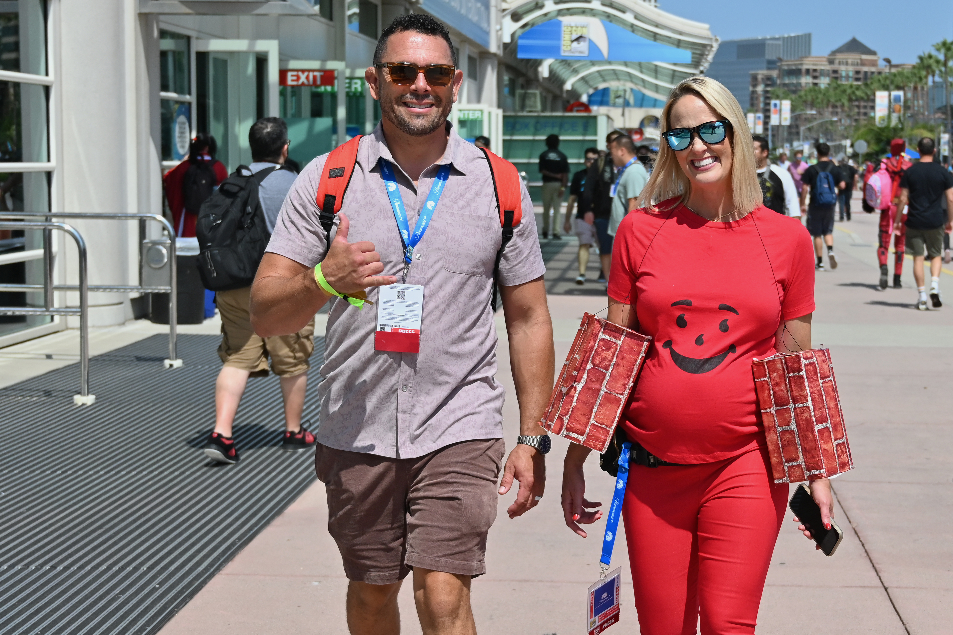A man and a pregnant woman walk outside a conference center. The woman wears a Kool-Aid Man costume with attached brick wall sleeves. The man gives a thumbs up