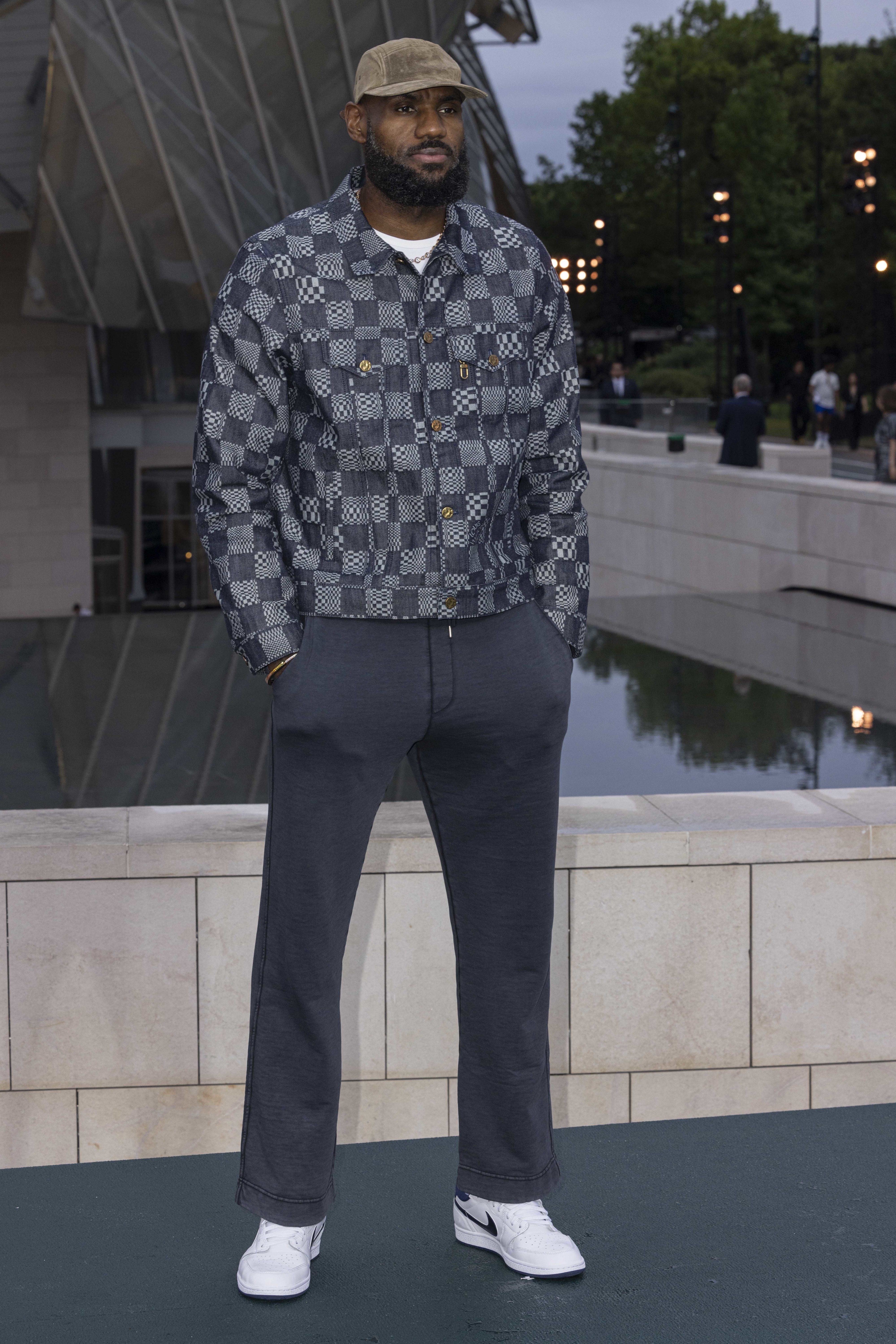 LeBron James stands outdoors wearing a patterned jacket, casual pants, a cap, and white sneakers