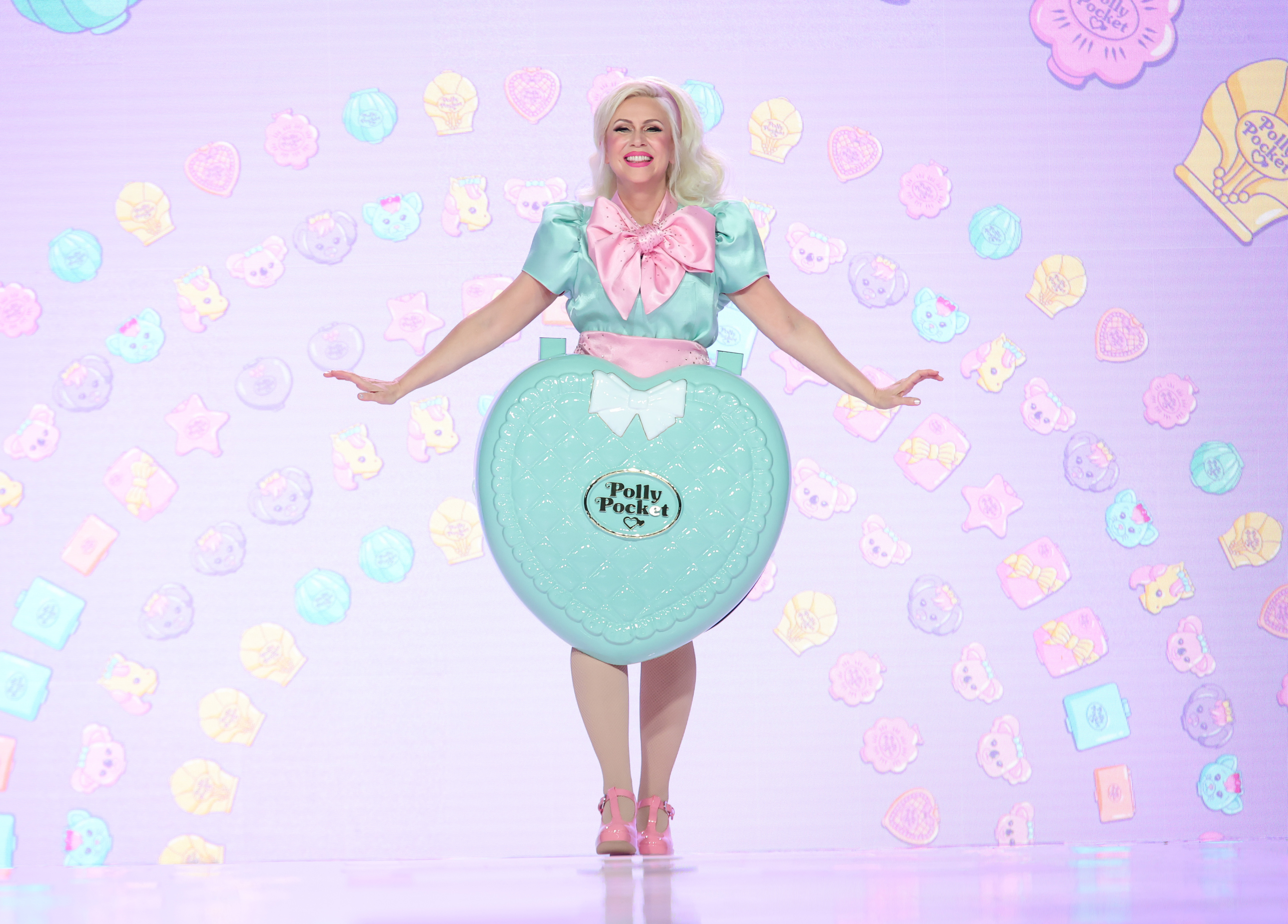 Woman dressed in a Polly Pocket-themed costume composed of a large heart-shaped accessory, light dress, and pink bow poses playfully on stage