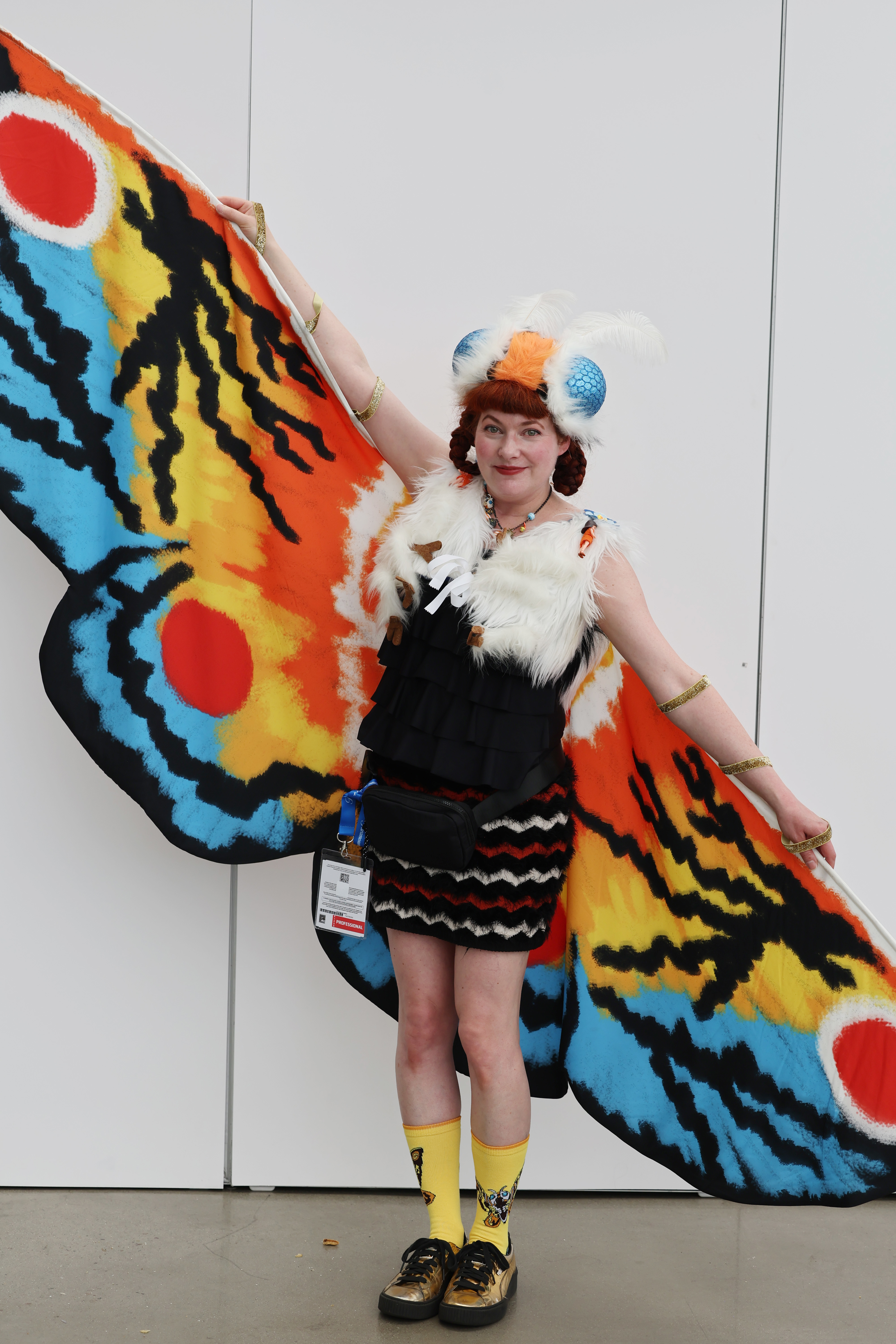 A person dressed in a colorful moth costume with large wings displays the costume by extending their arms. The person is wearing a fur-trimmed top, tiered skirt, and yellow socks with metallic shoes