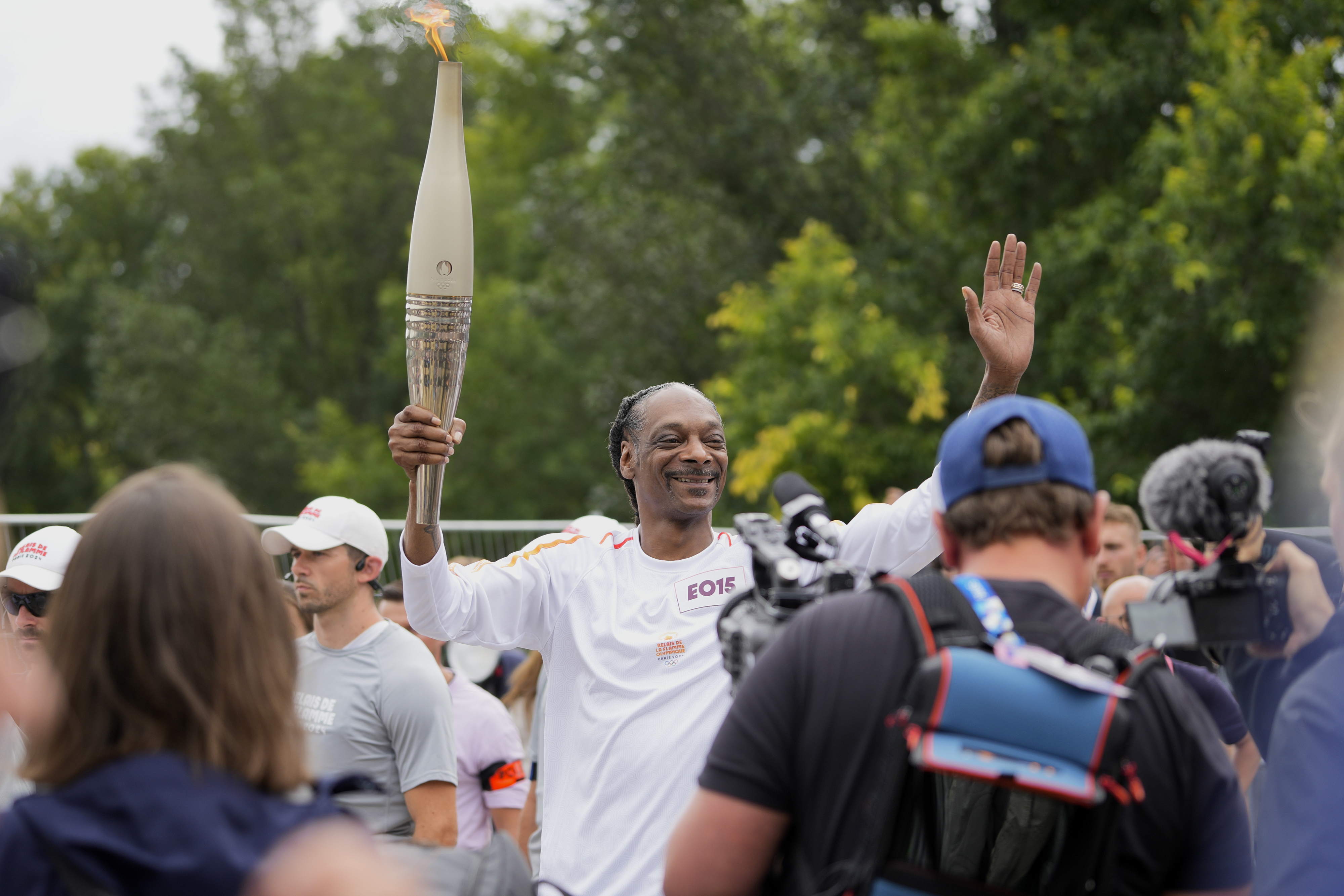 Snoop Dogg smiling and waving while holding a large torch at an outdoor event, surrounded by people and camera operators