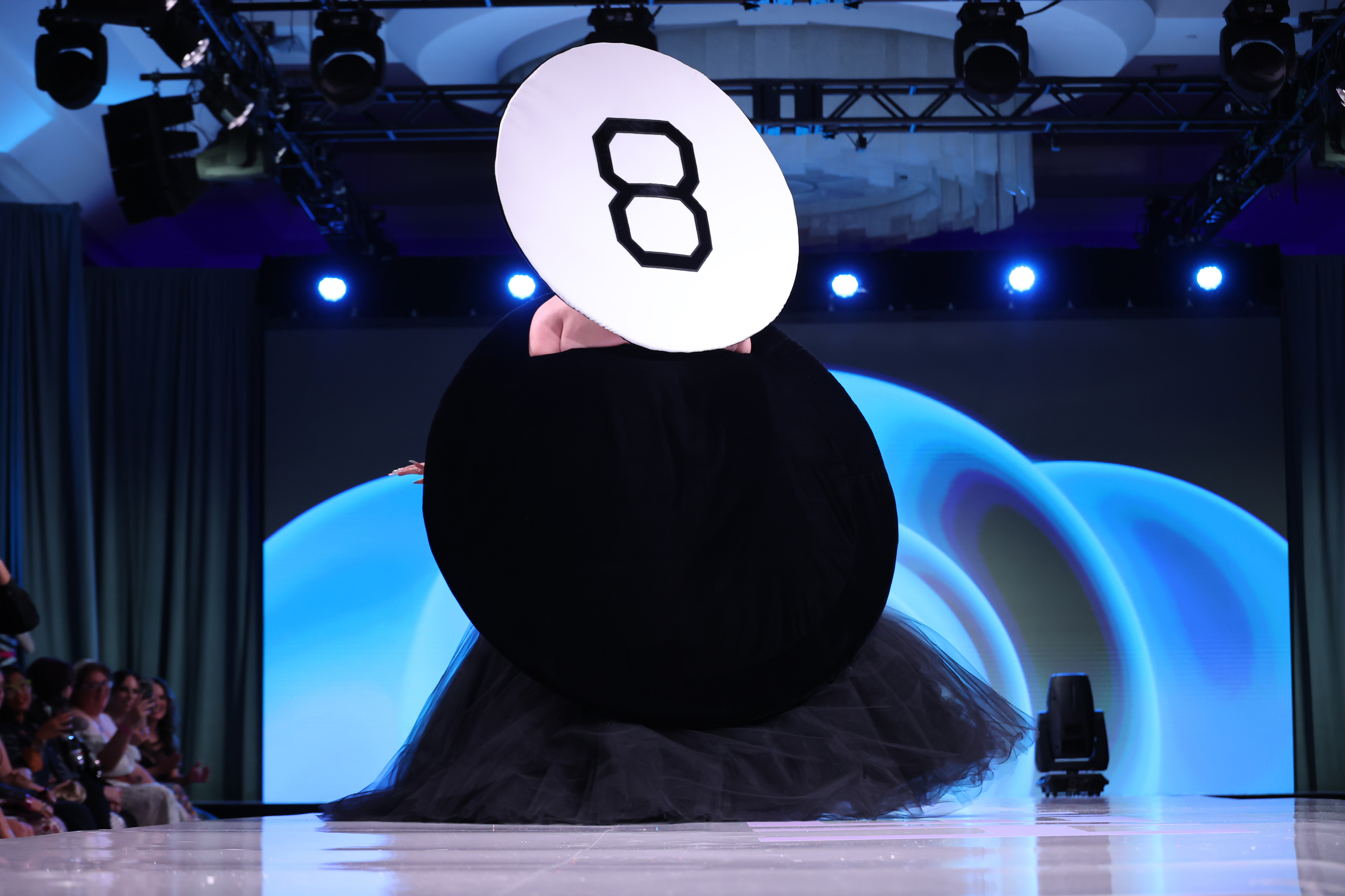 Person wearing a dress resembling a large black pool eight ball on a runway, with the number 8 prominently displayed