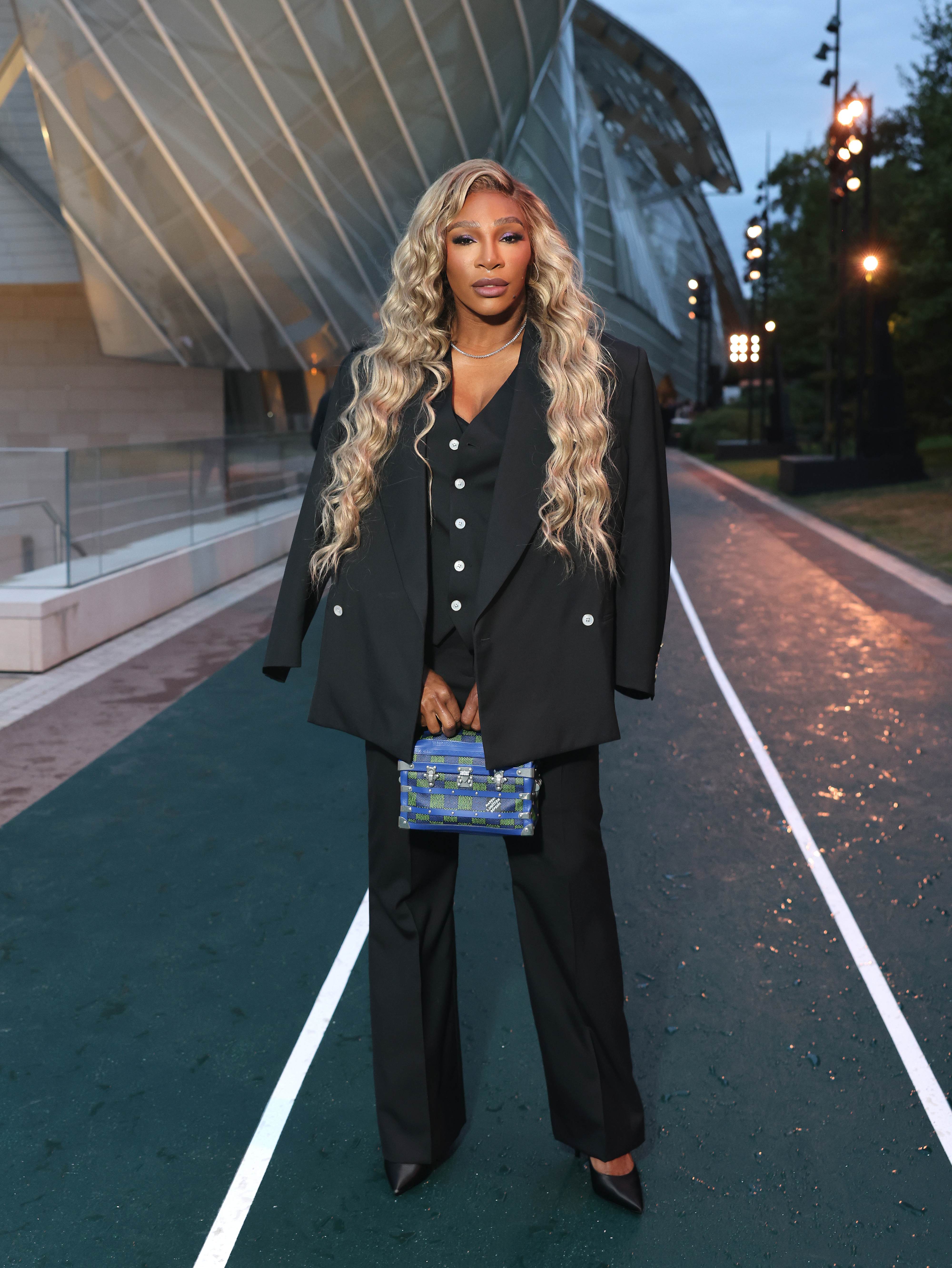 Serena Williams stands outdoors in an oversized black suit, wearing a long blonde wig and holding a small patterned bag
