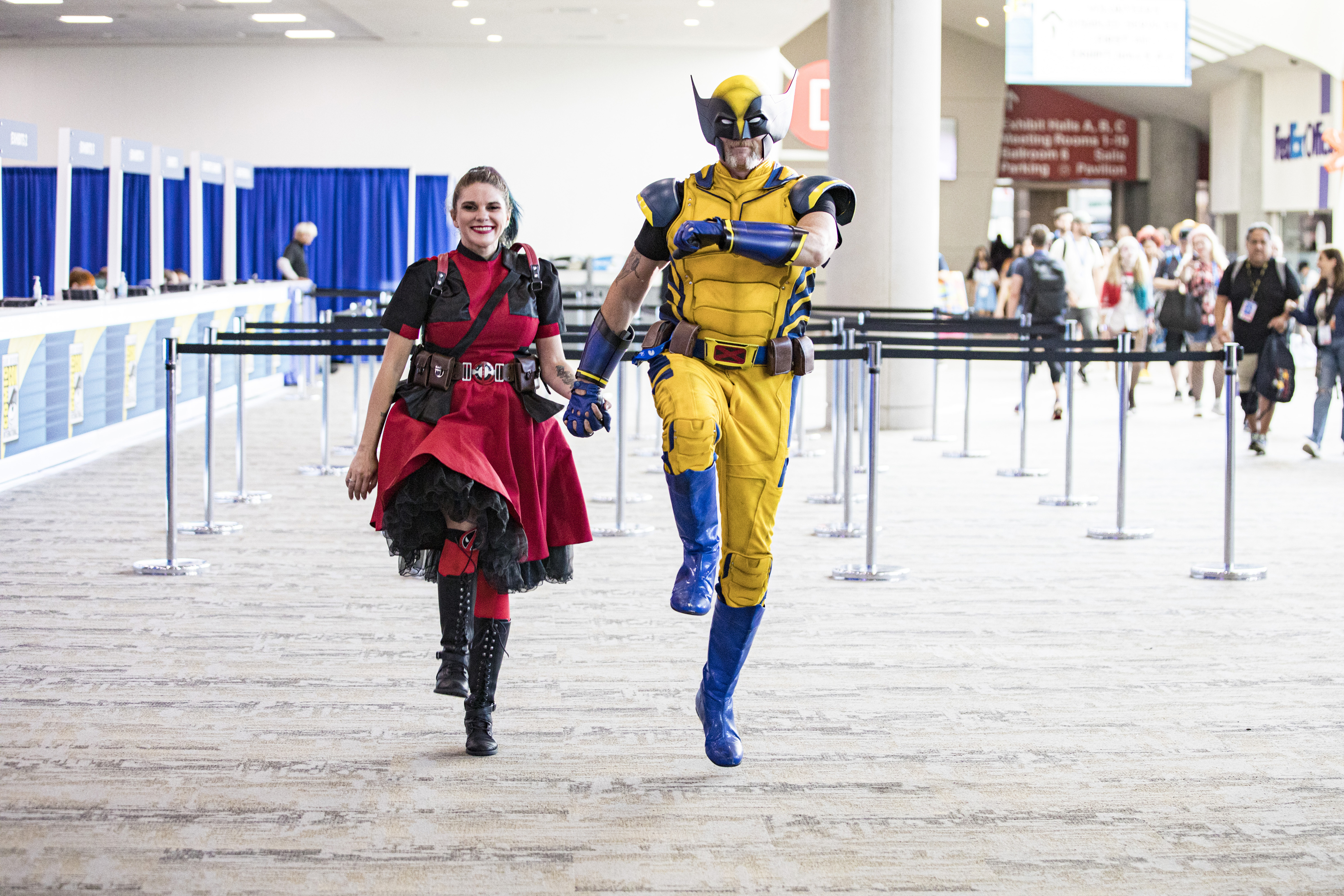 Cosplayers dressed as Scarlet Witch and Wolverine walk energetically down a convention hallway, with booths and attendees visible in the background
