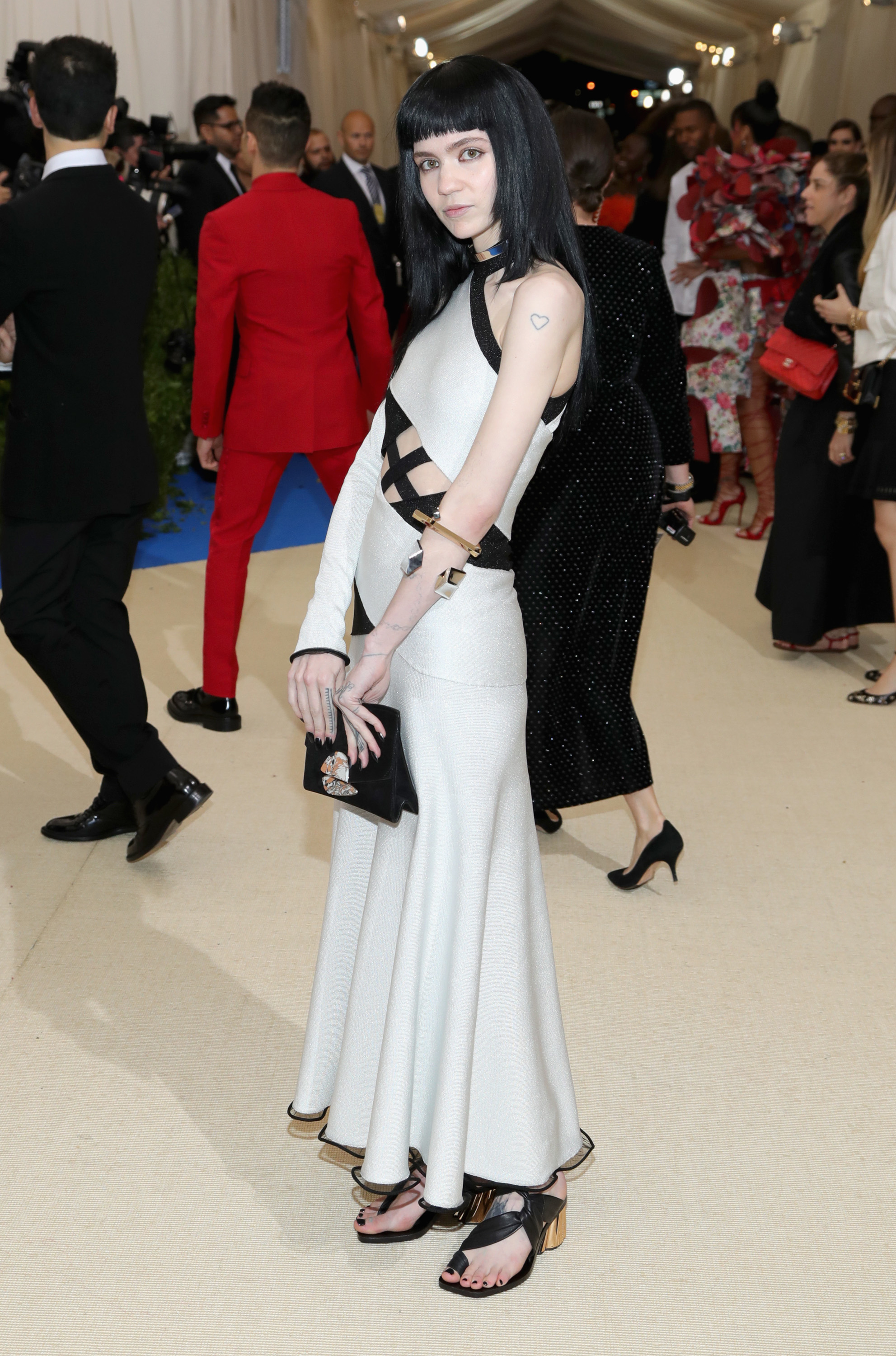 Grimes on the red carpet wearing a sleeveless, white gown with black cut-out details, holding a black purse. She has shoulder-length black hair