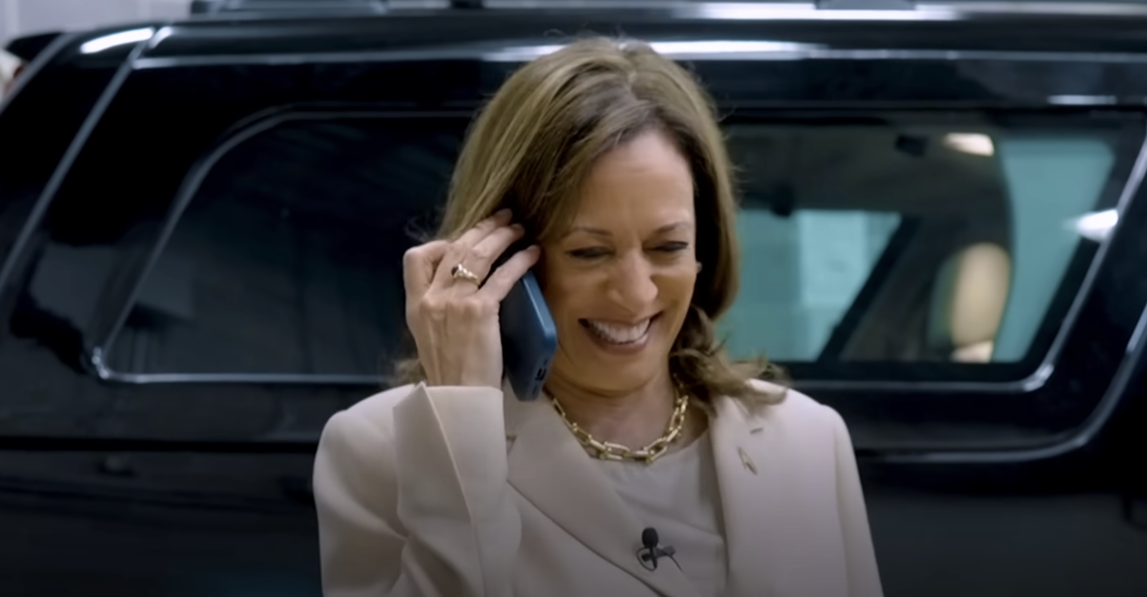 Kamala Harris, wearing a beige suit and a gold chain necklace, smiles while talking on a cell phone. She stands in front of a vehicle
