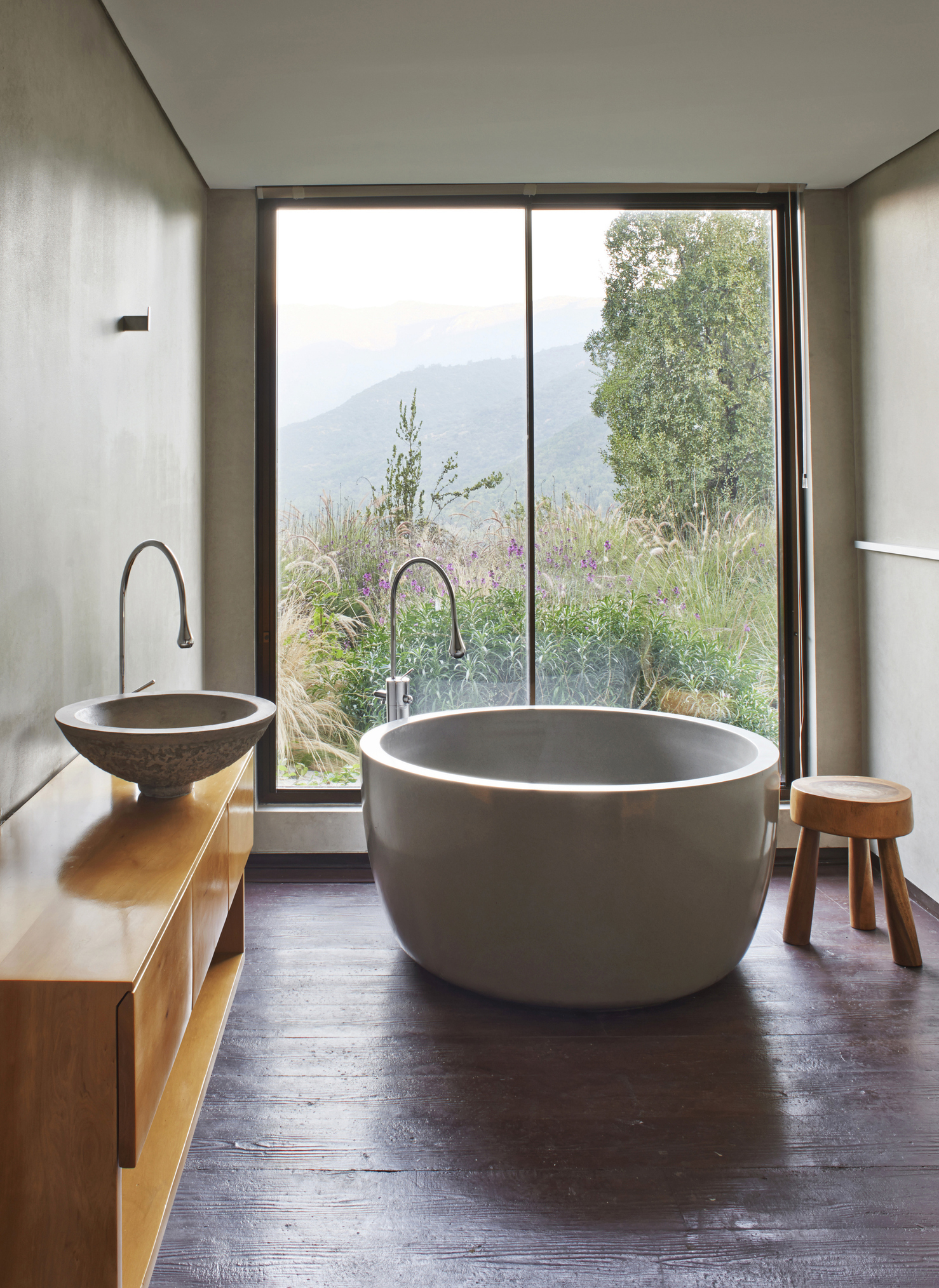 Modern bathroom features a freestanding bathtub, vessel sink on a wooden vanity, small stool, and a large window with an outdoor scenic view