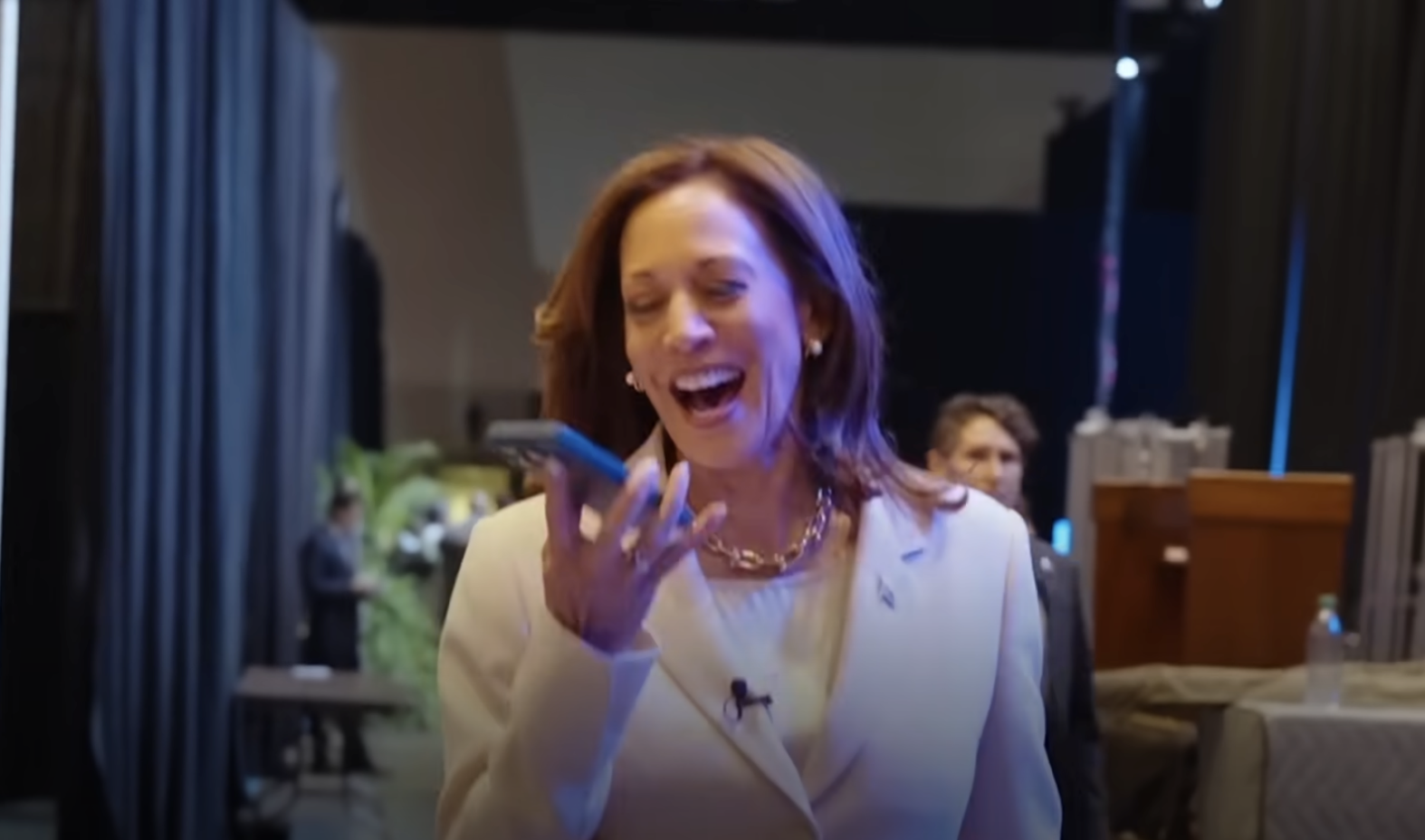 Kamala Harris is smiling and looking at her phone, wearing a business suit with a necklace. She is indoors, walking in a dimly-lit area