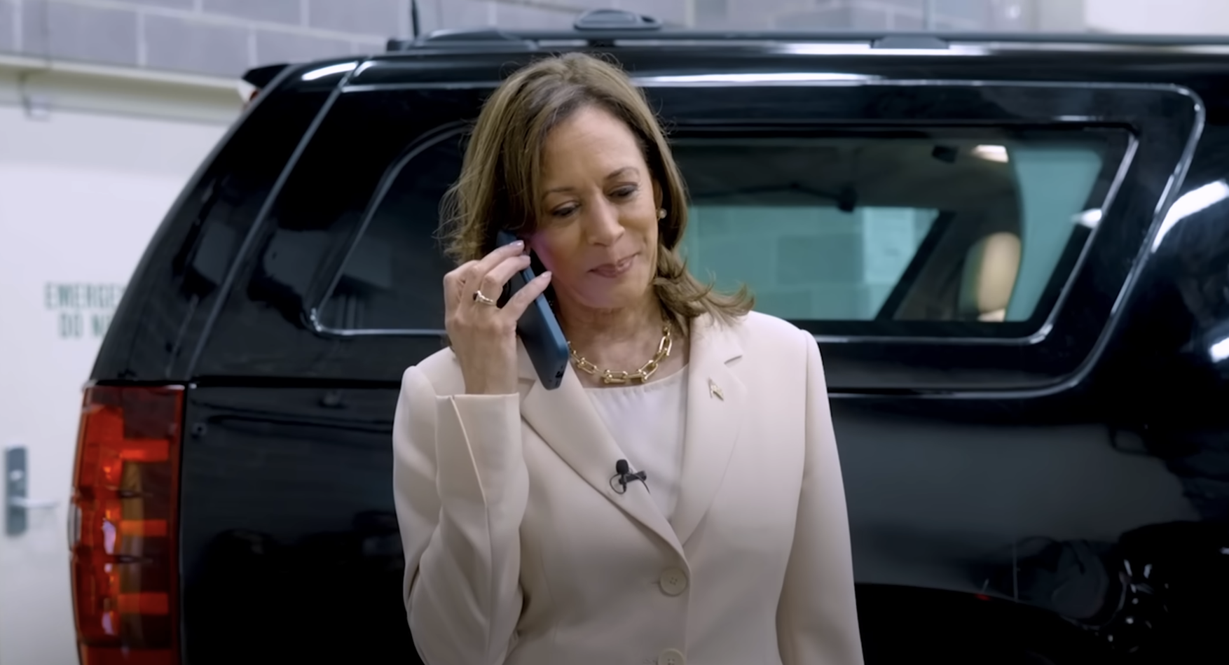 Kamala Harris stands next to a vehicle, smiling and holding a phone to her ear, wearing a beige blazer