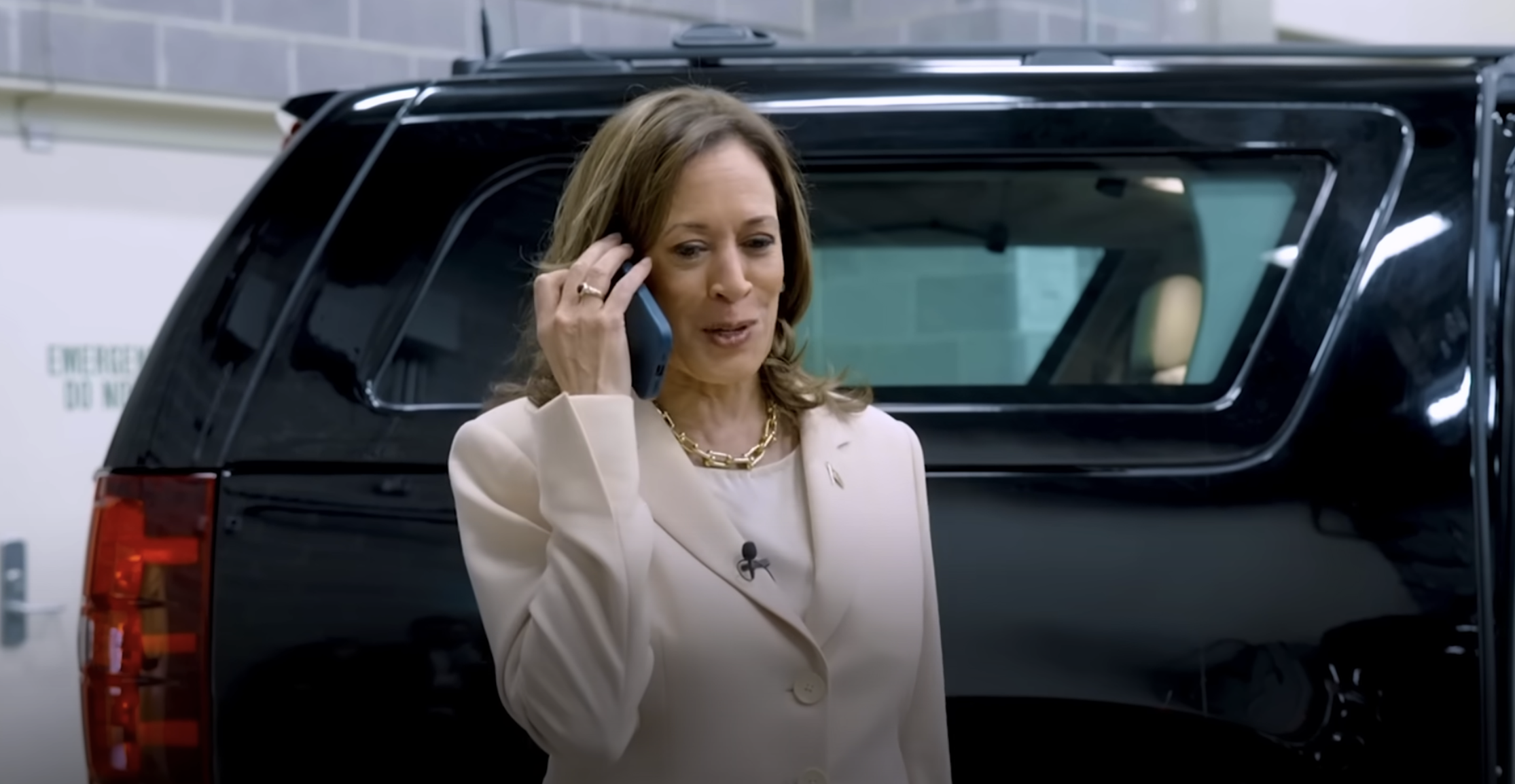 Kamala Harris is talking on a smartphone, smiling, wearing a light-colored suit, standing near a black SUV in a garage