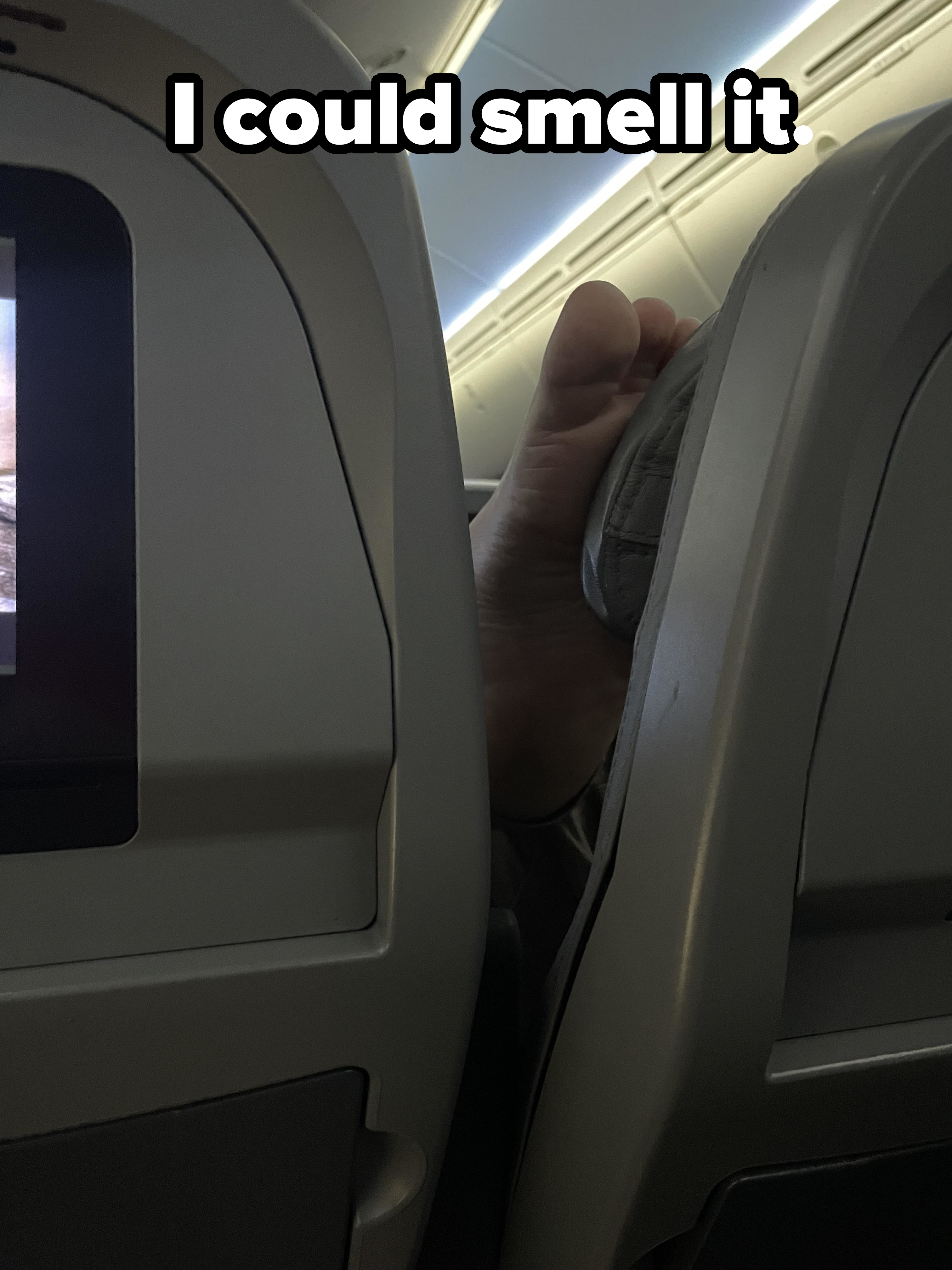 A bare foot is resting on top of the armrest of an airline seat, intruding into the personal space of the passenger seated behind